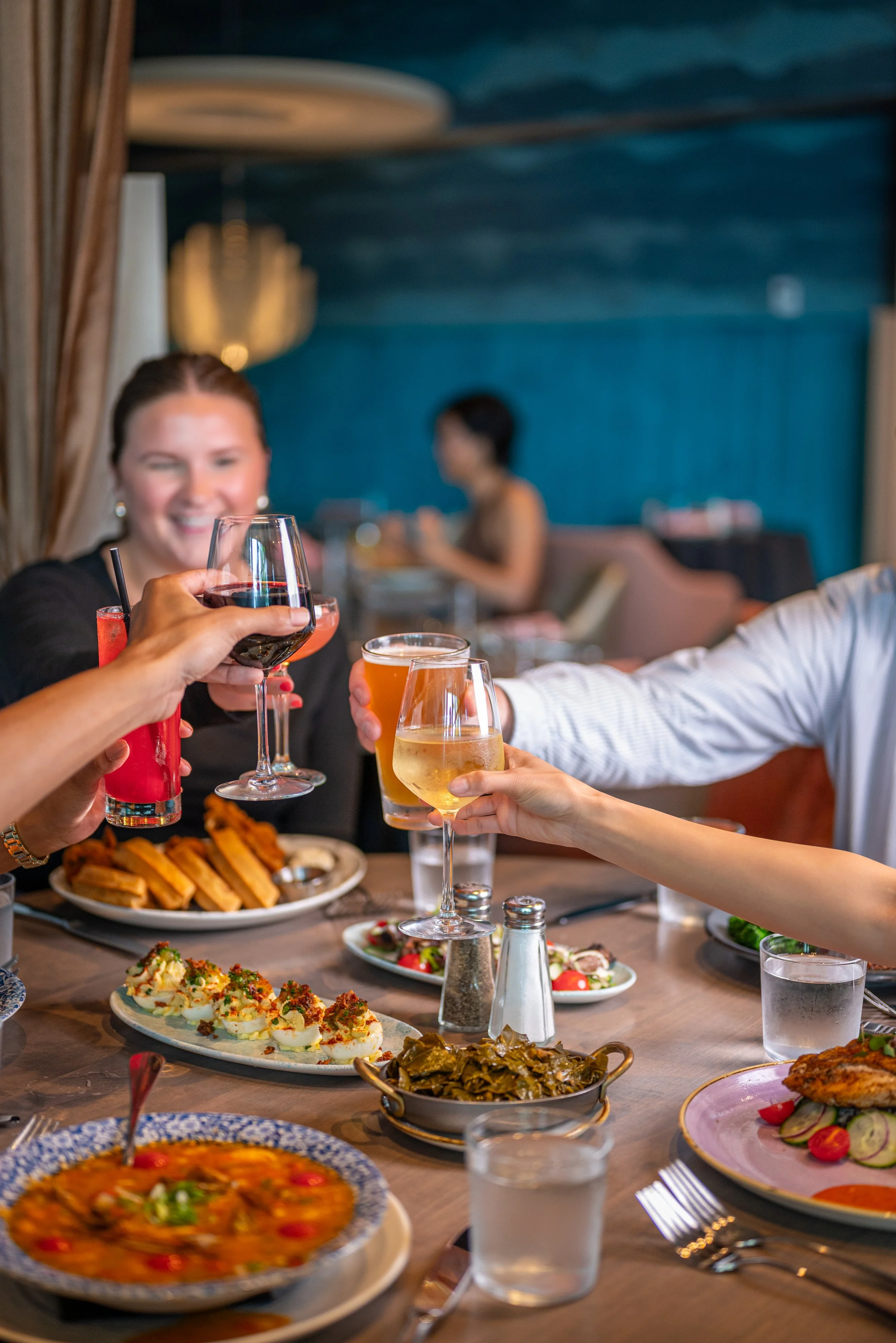 Hands clinking glasses of wine, beer, and cocktails over a table filled with plated dishes and sides in a restaurant setting.