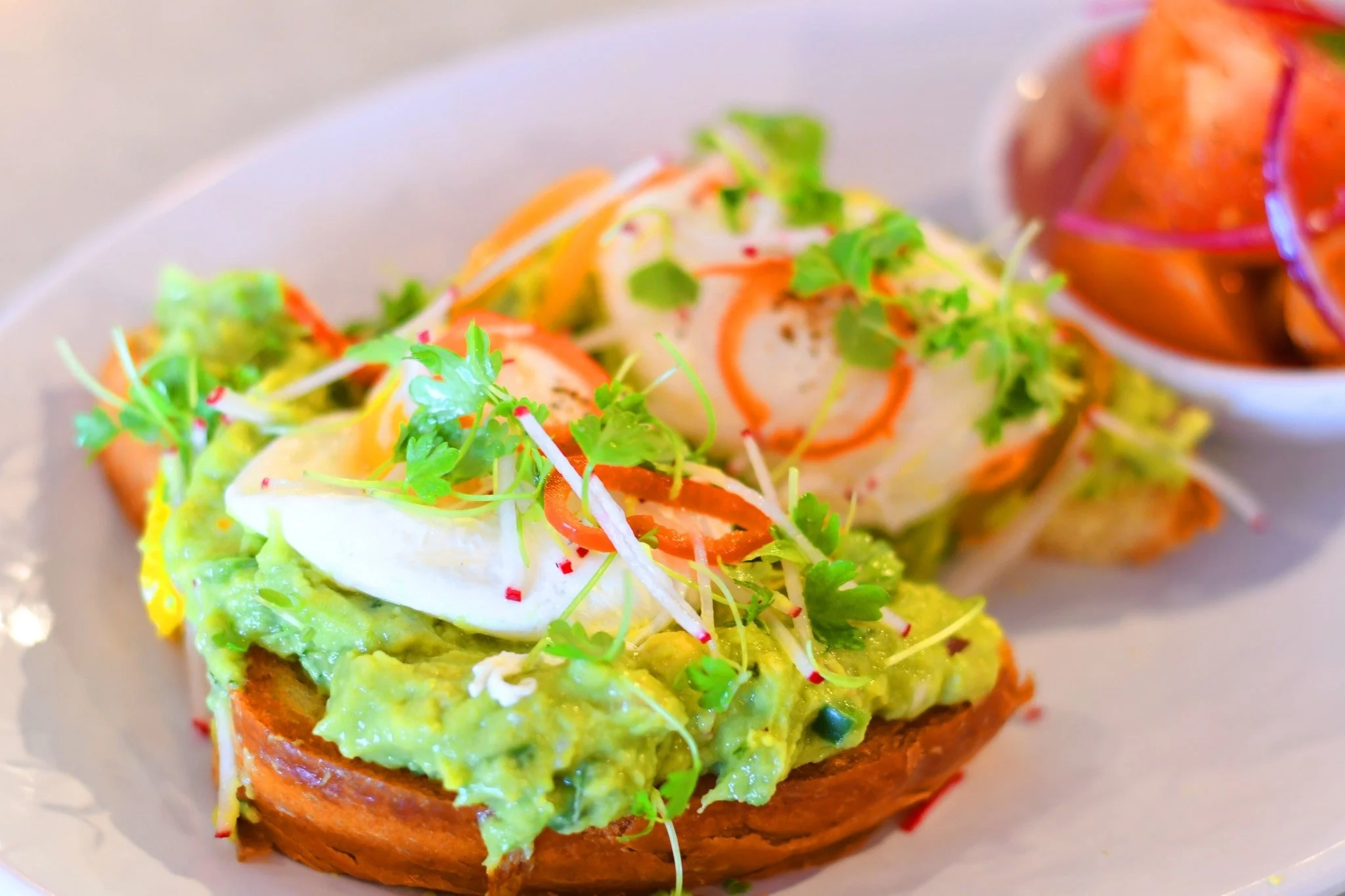 Avocado toast topped with poached eggs, microgreens, and sliced radishes on a white plate, with a side of sliced fruit and onions in a small bowl.