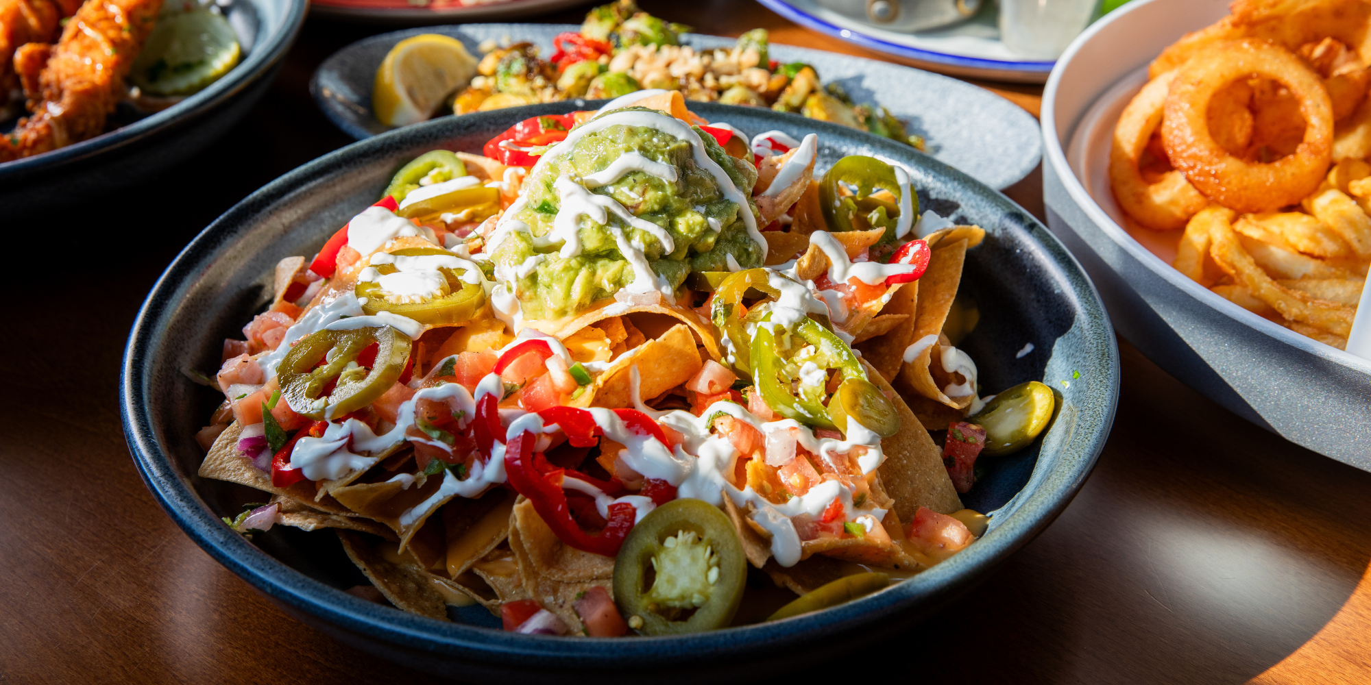 Loaded nachos topped with cheese, jalapeños, tomatoes, sour cream, and guacamole, served with other appetizers and sides on a restaurant table.