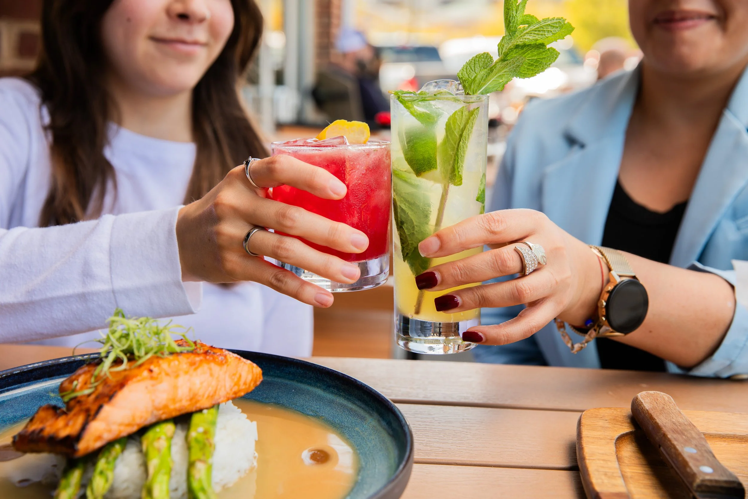 Two people clinking a red cocktail and a tall mojito over a plate of grilled salmon with asparagus and rice on a wooden table.