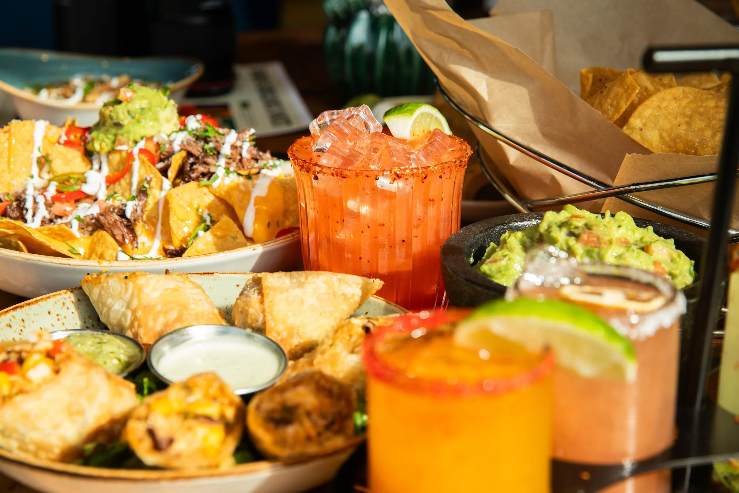 Assorted Mexican appetizers including nachos, guacamole, egg rolls, and colorful cocktails on a wooden table.