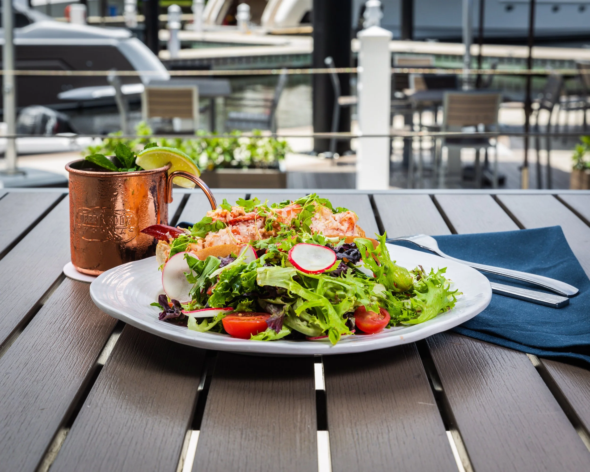 A plate of lobster roll with mixed greens, cherry tomatoes, and radish slices on a wooden table, served with a copper mug cocktail garnished with lime and mint.