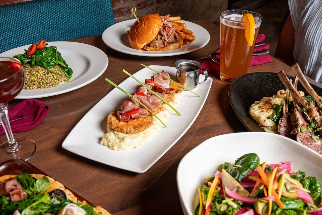 A table with assorted dishes including fried green tomatoes with prosciutto, a burger with fries, lamb chops, salads, and drinks including beer and cocktails.
