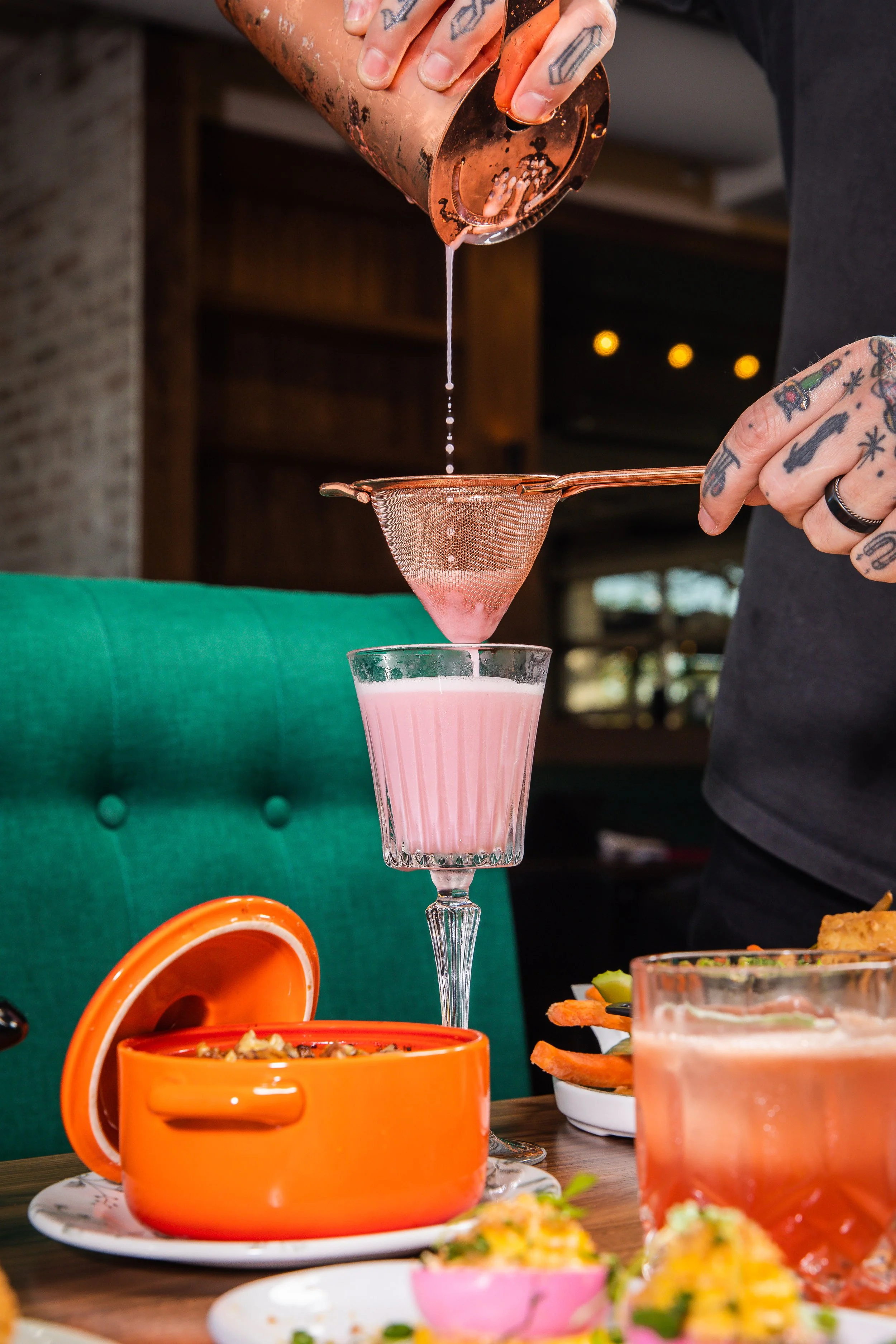 A bartender straining a pink cocktail into a tall glass over a table with colorful dishes, including an orange pot and garnished plates.