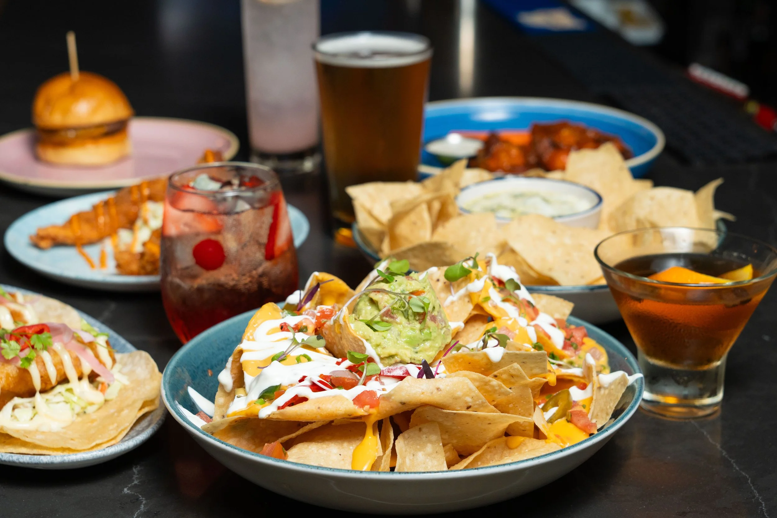A table with nachos topped with guacamole and sour cream, tacos, a slider, wings, and assorted drinks including beer and cocktails.