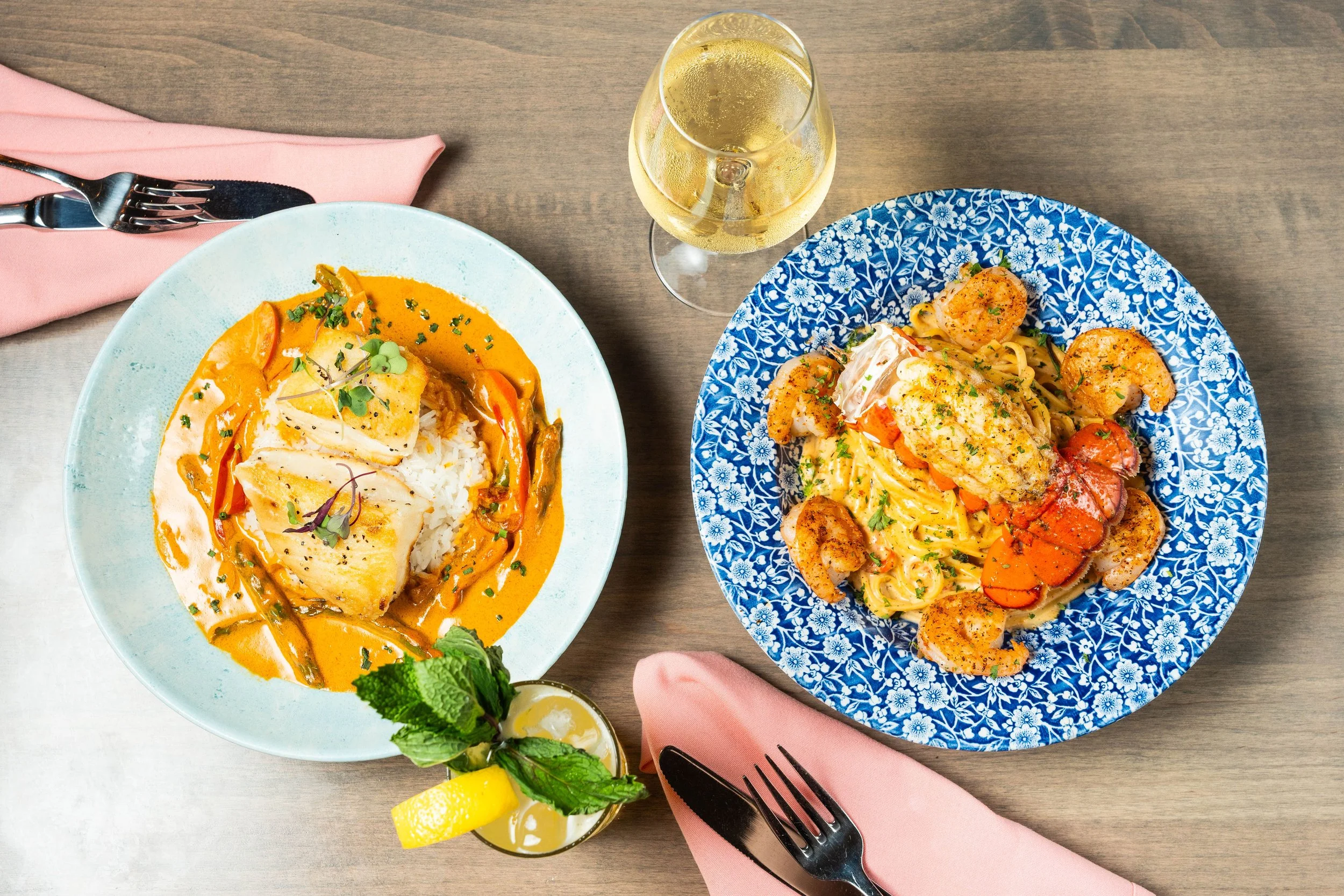Two plates of seafood dishes on a wooden table: one with fish in orange sauce over rice, the other with lobster tail and shrimp over pasta, served with drinks and pink napkins.