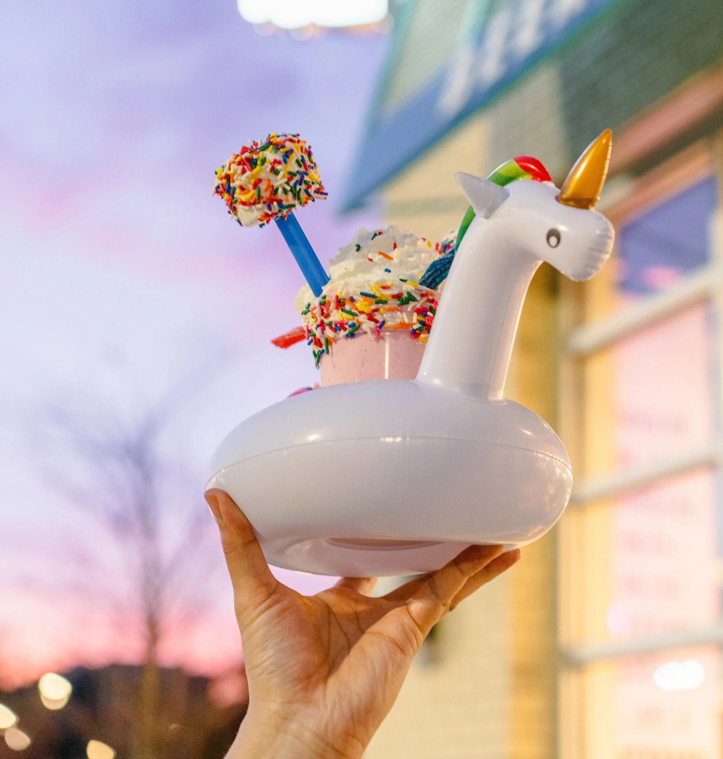 A hand holding a unicorn-shaped float with a cup of ice cream topped with sprinkles and colorful treats, against a sunset sky near a building.