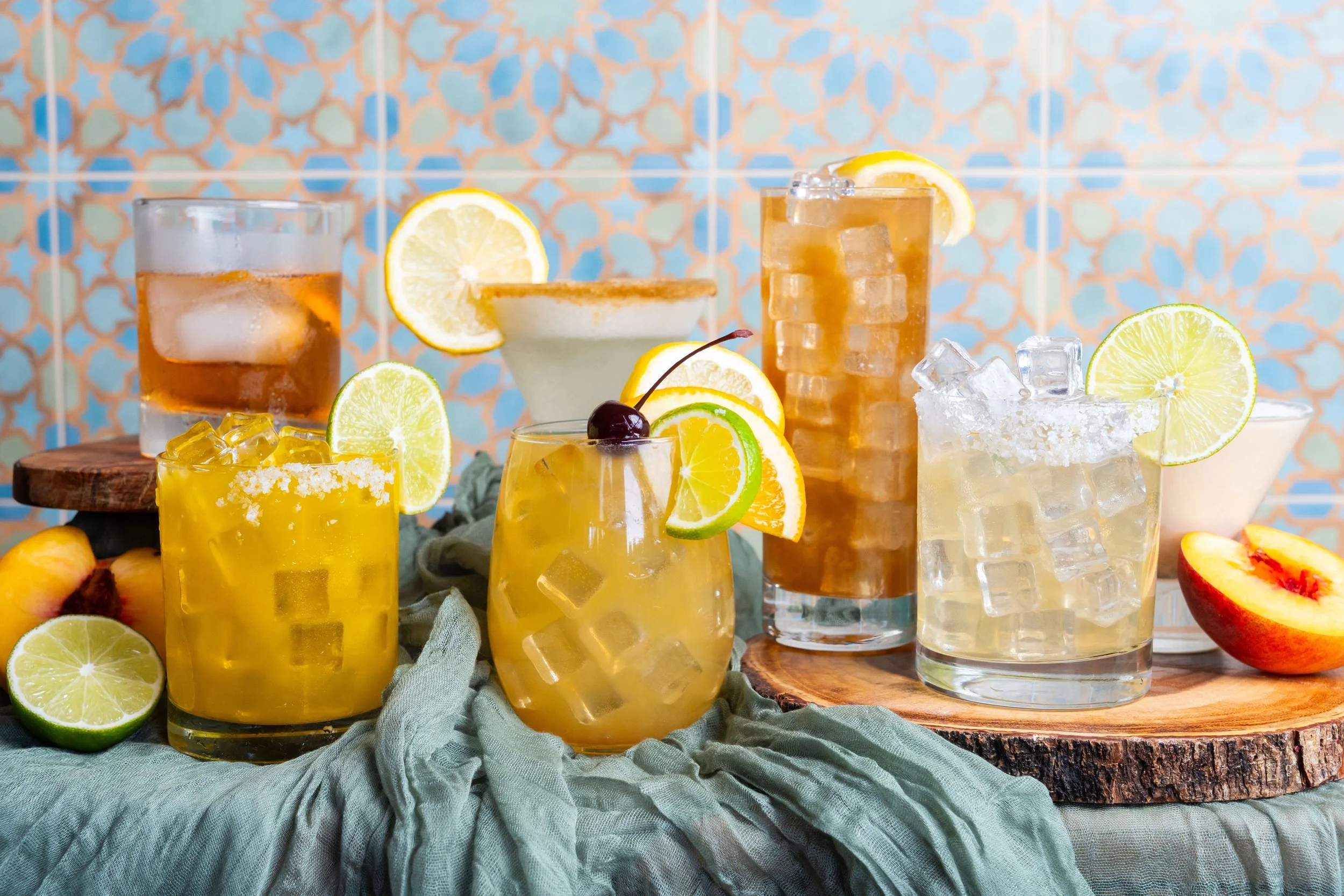 Six cocktails on a table with a green cloth, garnished with lime, lemon, and cherries, in front of a patterned tile backdrop with peaches and citrus fruits around.