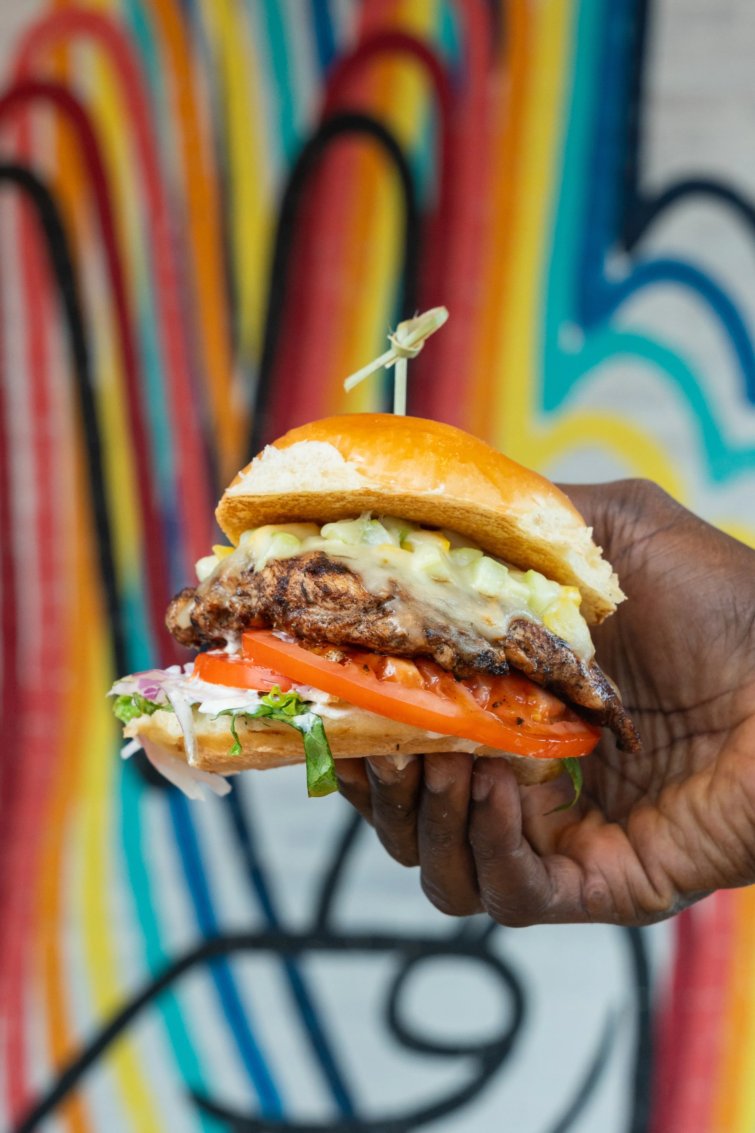 A hand holding a large burger with lettuce, tomato, melted cheese, and a toasted bun, against a colorful graffiti-style background.