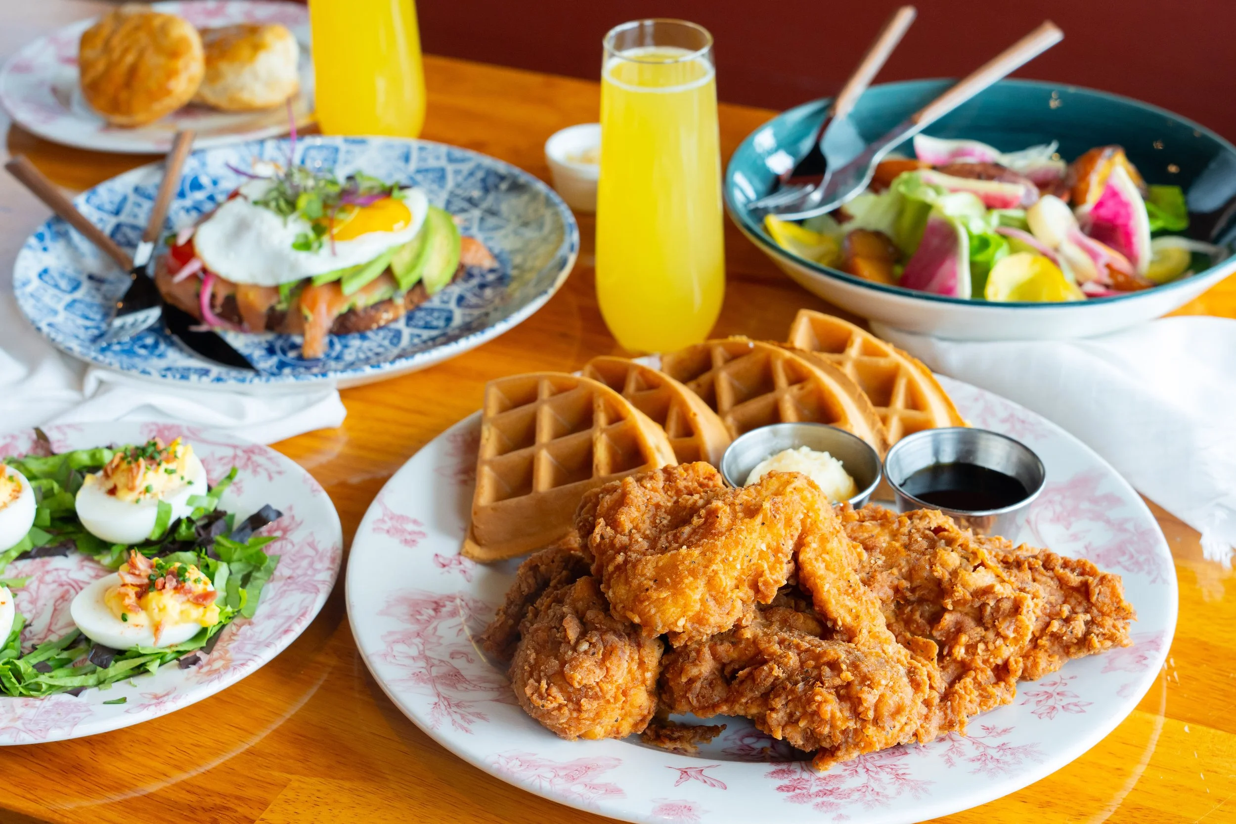 Plate of fried chicken and waffles with butter and syrup, surrounded by deviled eggs, salad, biscuits, and two glasses of orange drink on a wooden table.