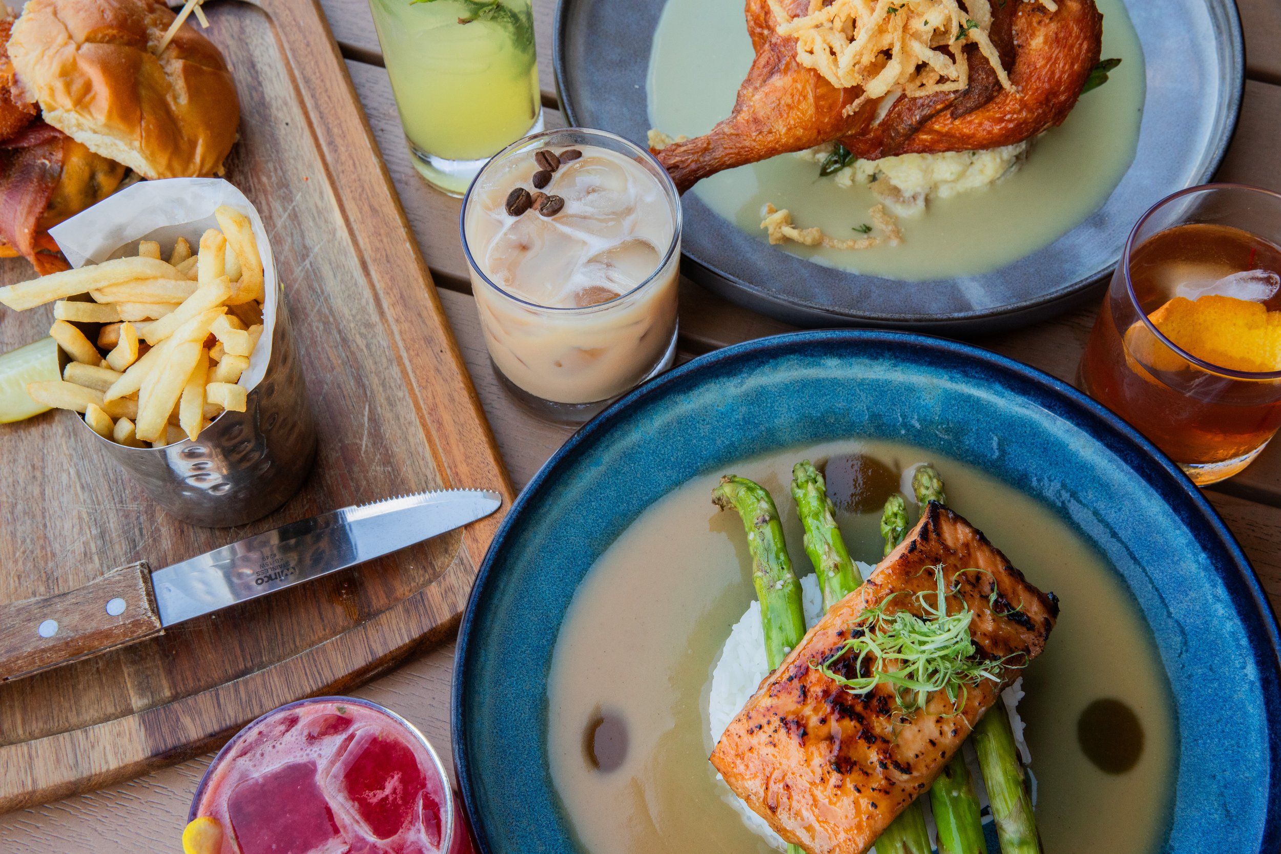 Table with salmon and asparagus in sauce, fried chicken on mashed potatoes, sliders with fries, and assorted cocktails.