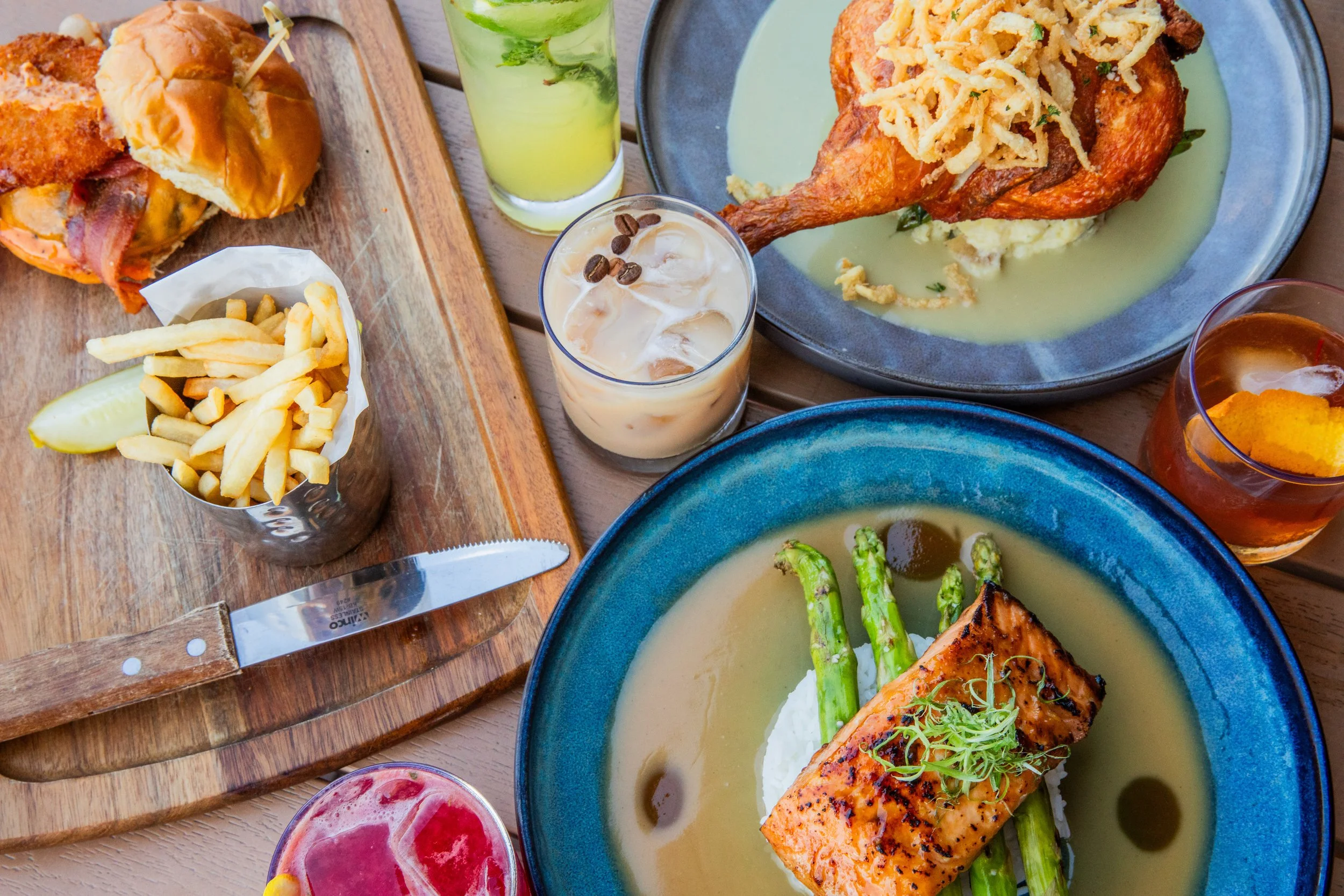 A table with plates of grilled salmon, fried chicken, fries, and assorted cocktails including mojito, iced coffee, and whiskey.