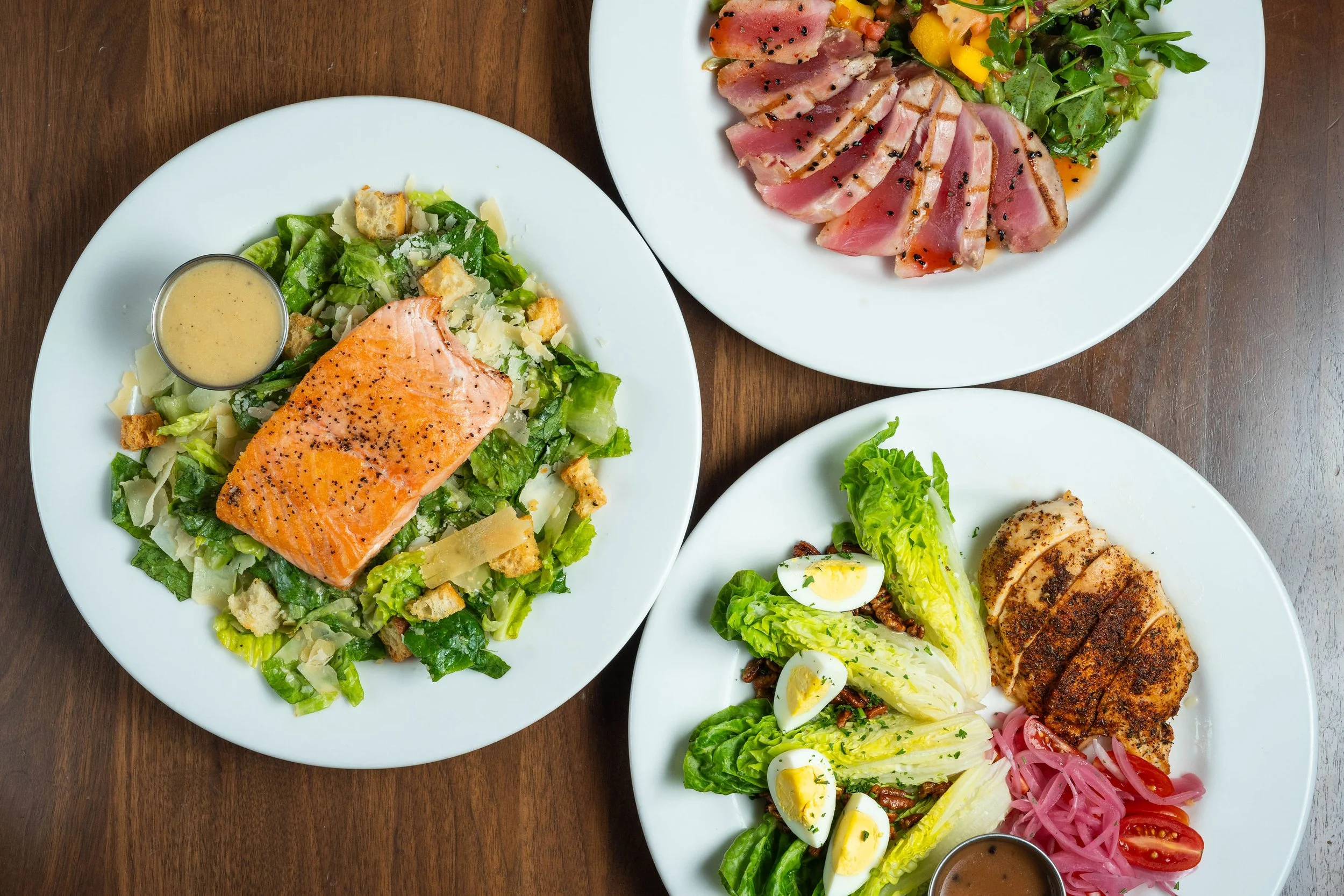 Three plates of food on a wooden table: salmon Caesar salad, seared tuna with greens, and a Cobb salad with chicken, eggs, and pickled onions.