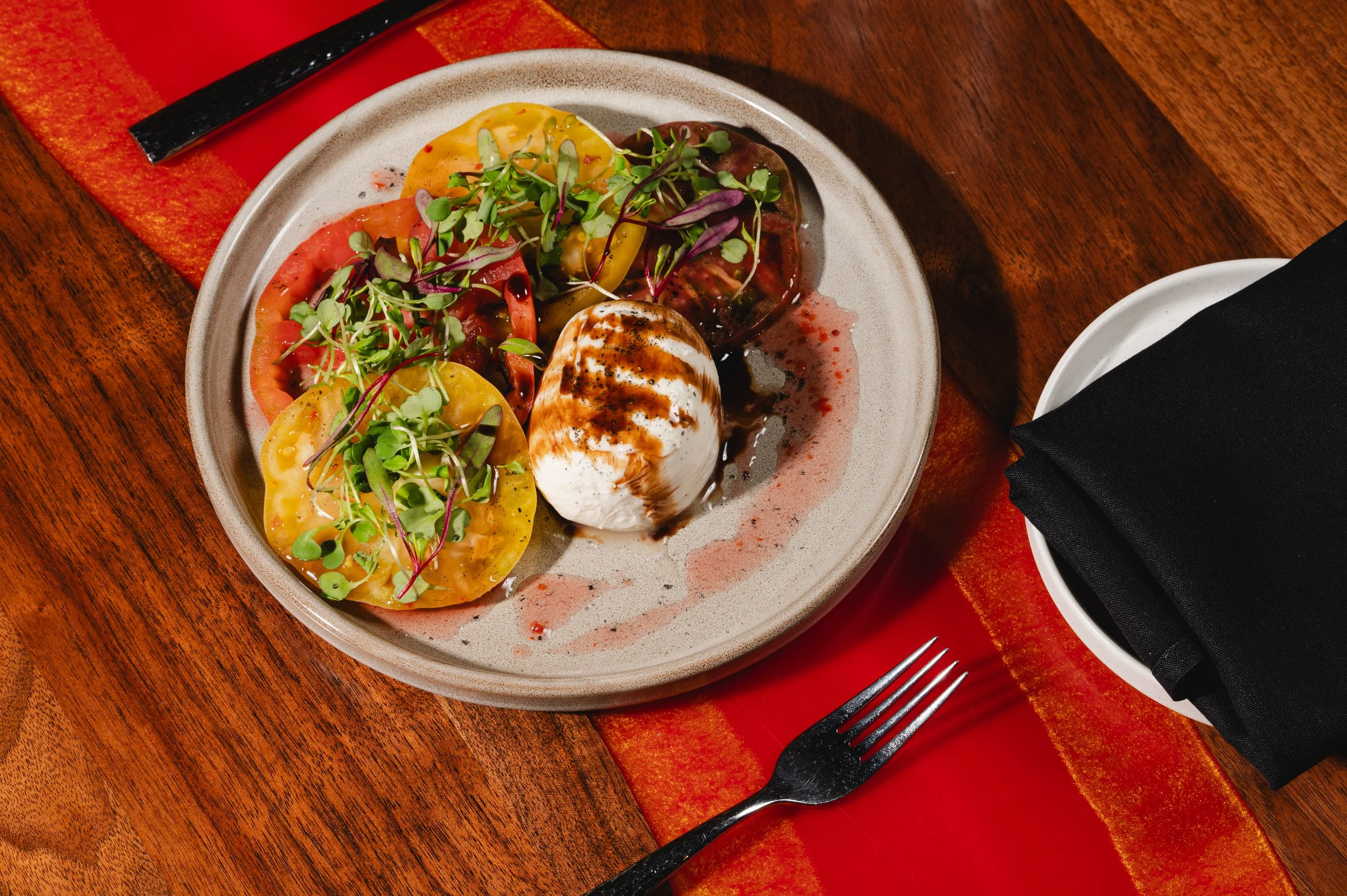 A plate of heirloom tomato slices topped with microgreens and balsamic glaze, served with a ball of fresh mozzarella on a wooden table with a red runner.