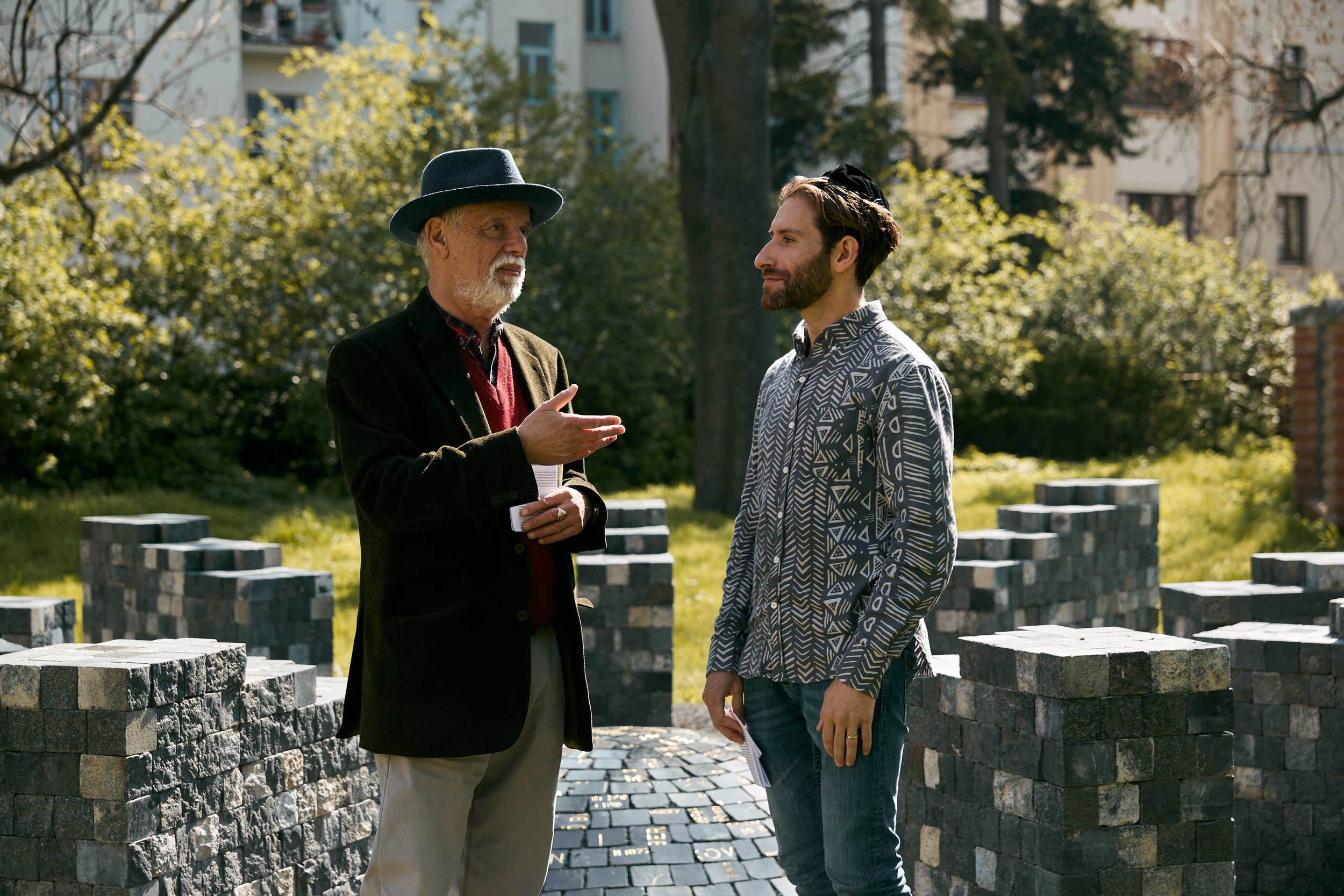 Two men are standing outdoors in a park, engaged in conversation. The older man on the left is wearing a dark blazer, a red sweater, and a gray hat. The younger man on the right has a beard, and is wearing a patterned button-up shirt and a black kippah. They are surrounded by greenery and modern buildings in the background.