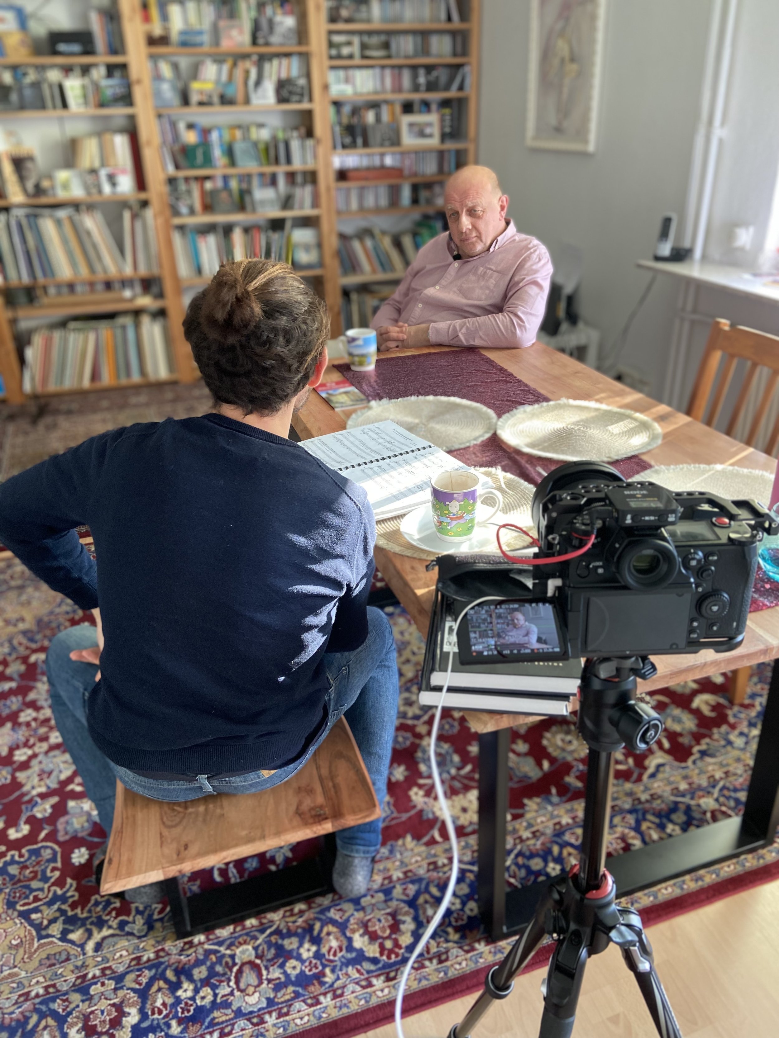 A woman conducting an interview with a bald man at a wooden dining table, with a camera recording the scene, set in a room with bookshelves and a patterned rug.