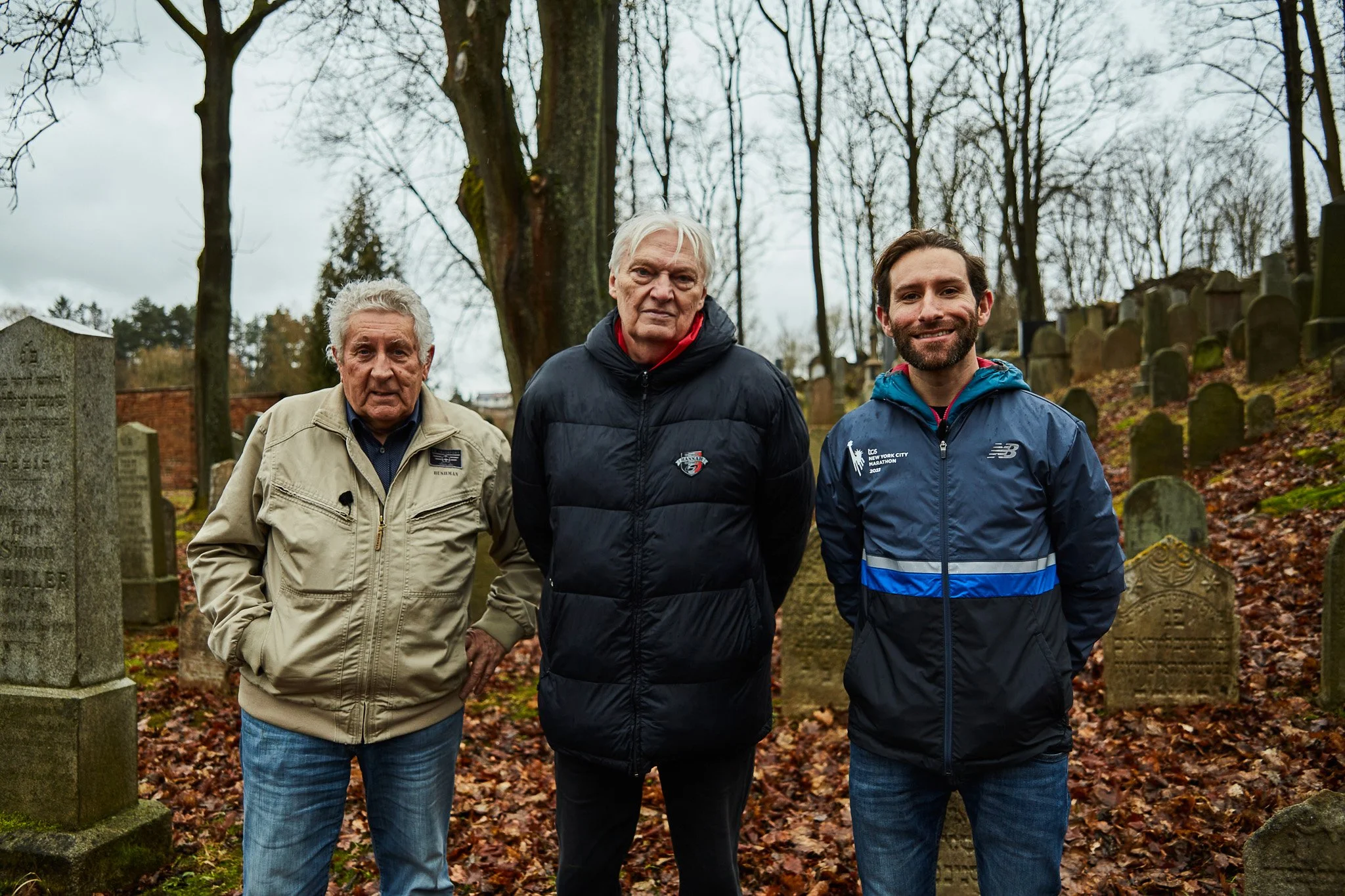 Three men standing in a cemetery with old gravestones and leafless trees in the background.