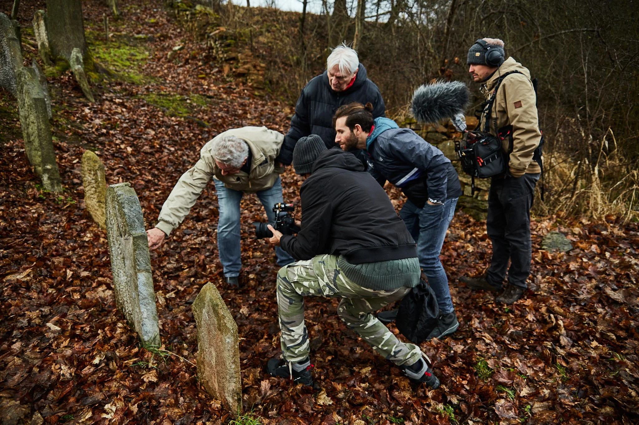 Group of five people gathered around a grave site outdoors in a wooded area on a rainy day, with leaves on the ground and bare trees in the background. They appear to be placing flowers or paying respects.