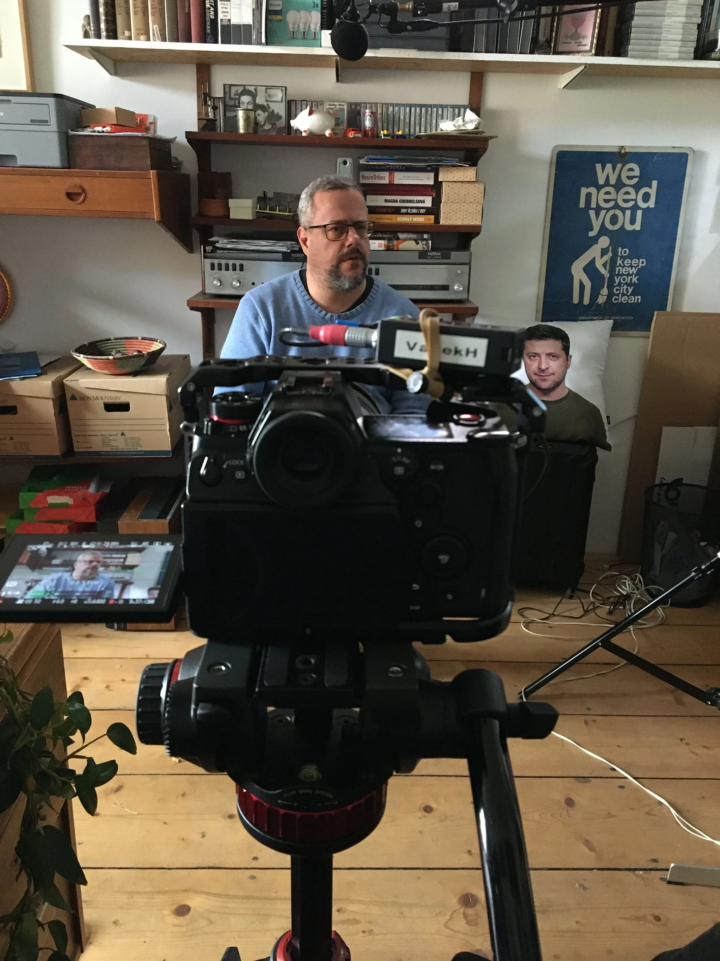 Man with glasses and a beard in a blue sweater in a room with bookshelves, framed pictures, and a blue sign that says 'we need you to keep new york city clean.' A professional camera on a tripod takes a photo of him, and the man's image appears on the camera's screen. There are various objects, boxes, and cords around the room.