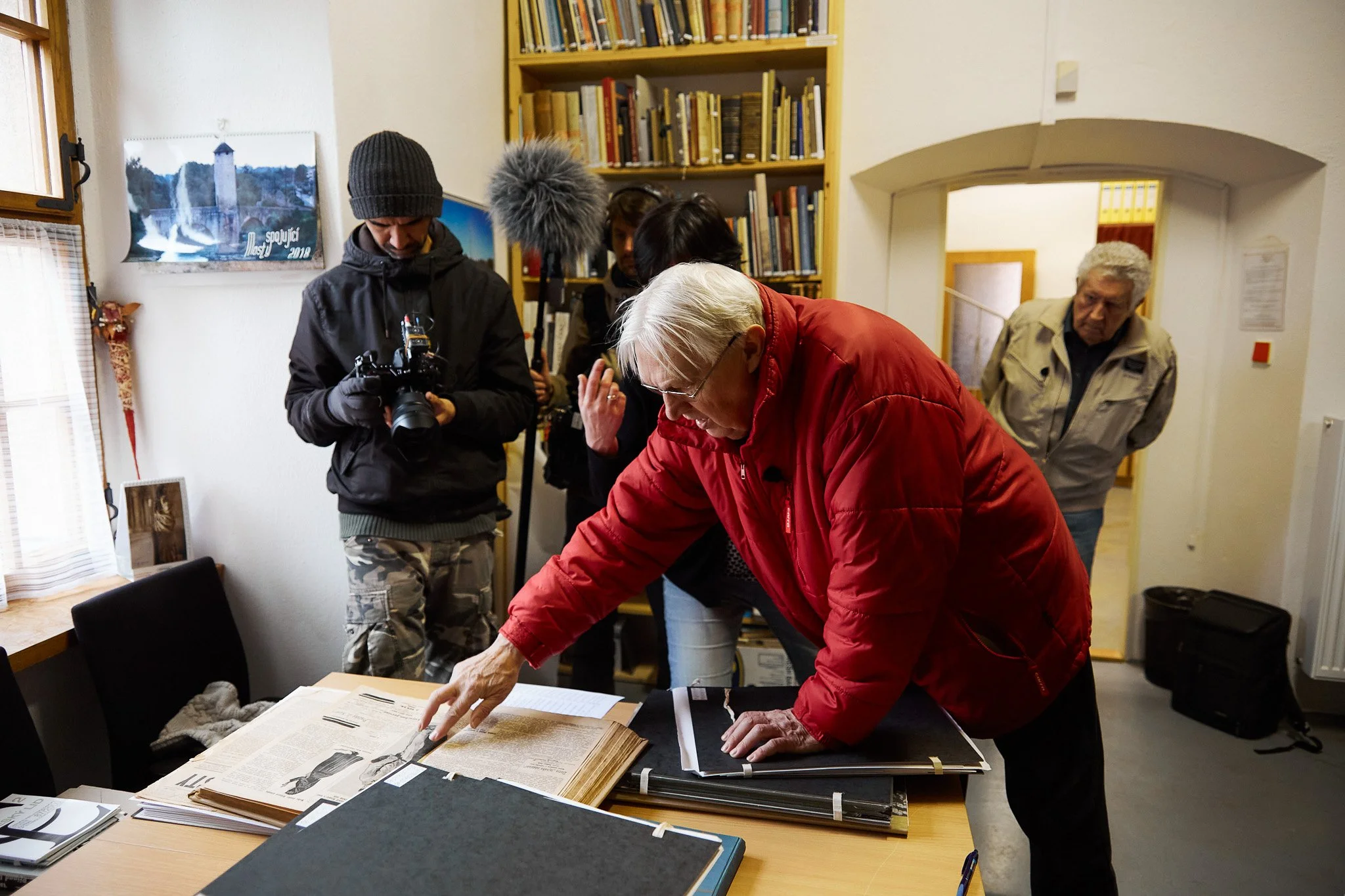 A group of people examining historical documents on a table in a room with bookshelves. One person is pointing at the documents, while others observe, with a photographer taking pictures.