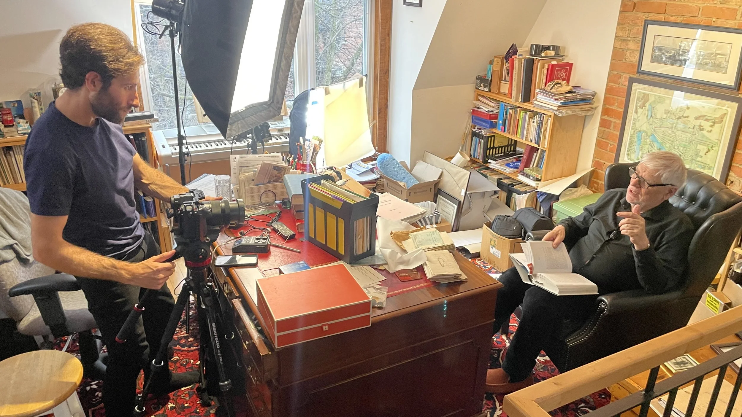 A man with a camera filming another man sitting in a leather armchair, reading a book, in a cluttered office with books, papers, and framed pictures on the wall.