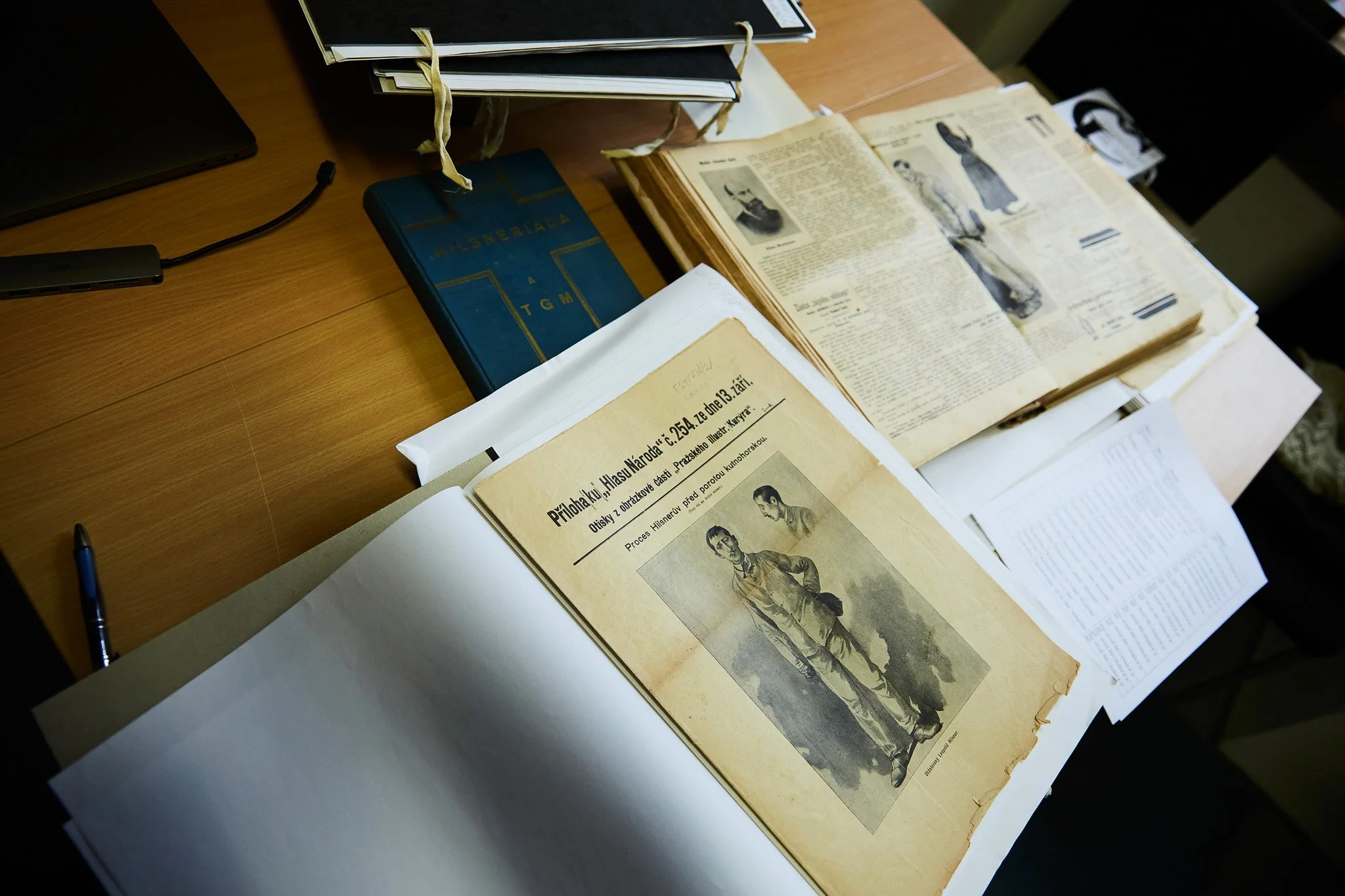 Historical documents and books spread out on a wooden desk, including a vintage newspaper with photographs and a blue hardcover book.