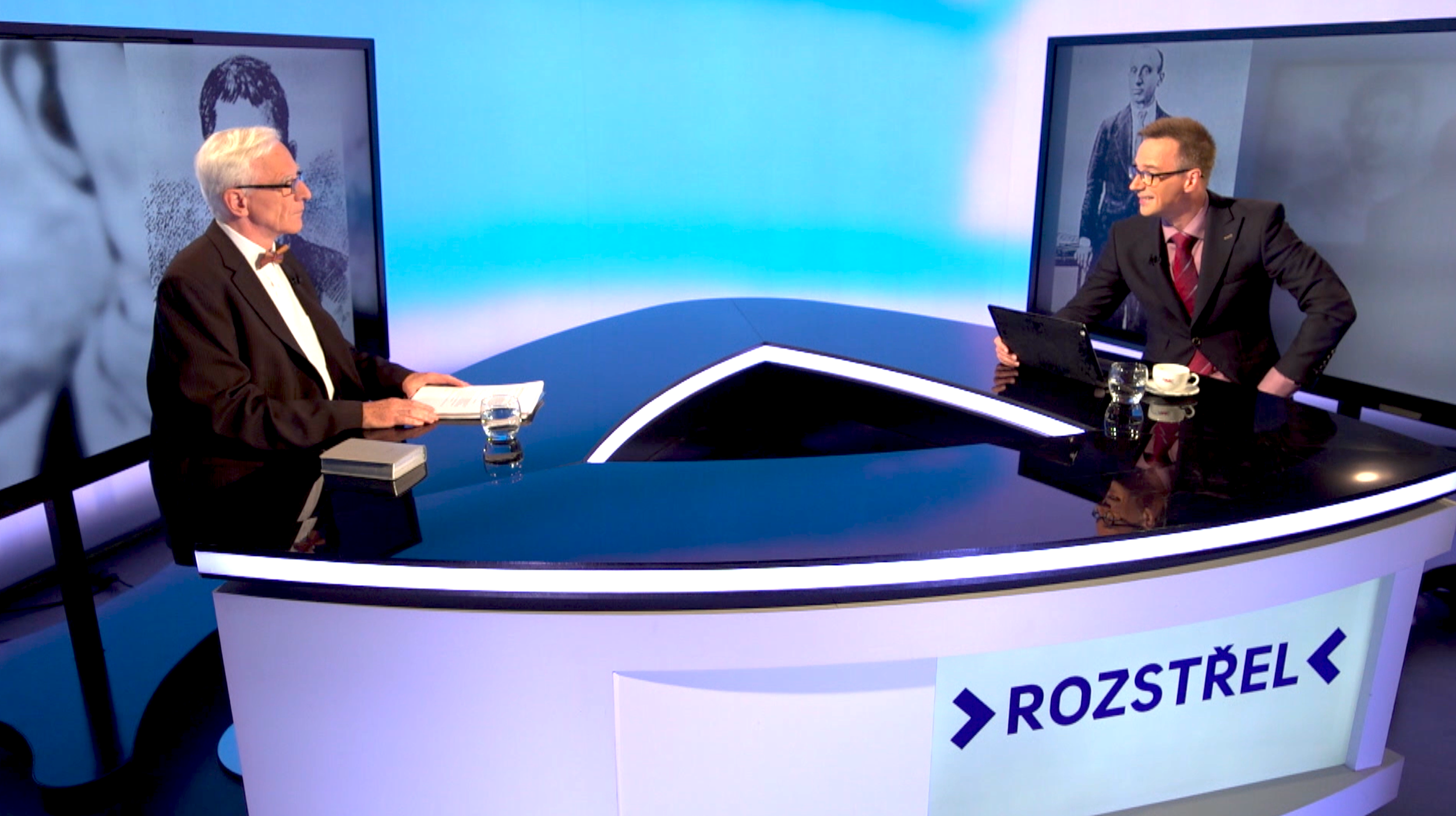 Two men in suits sit across from each other at a technological television studio desk. One man is elderly with white hair and glasses, wearing a black tuxedo with a bow tie, and the other is younger, with glasses and light brown hair, dressed in a dark suit with a red tie. Both have glasses and are engaged in conversation. There are large television screens behind them displaying black and white images. The studio has a modern design with a curved black desk with illuminated edges and a sign that reads 'ROZSTREL'.