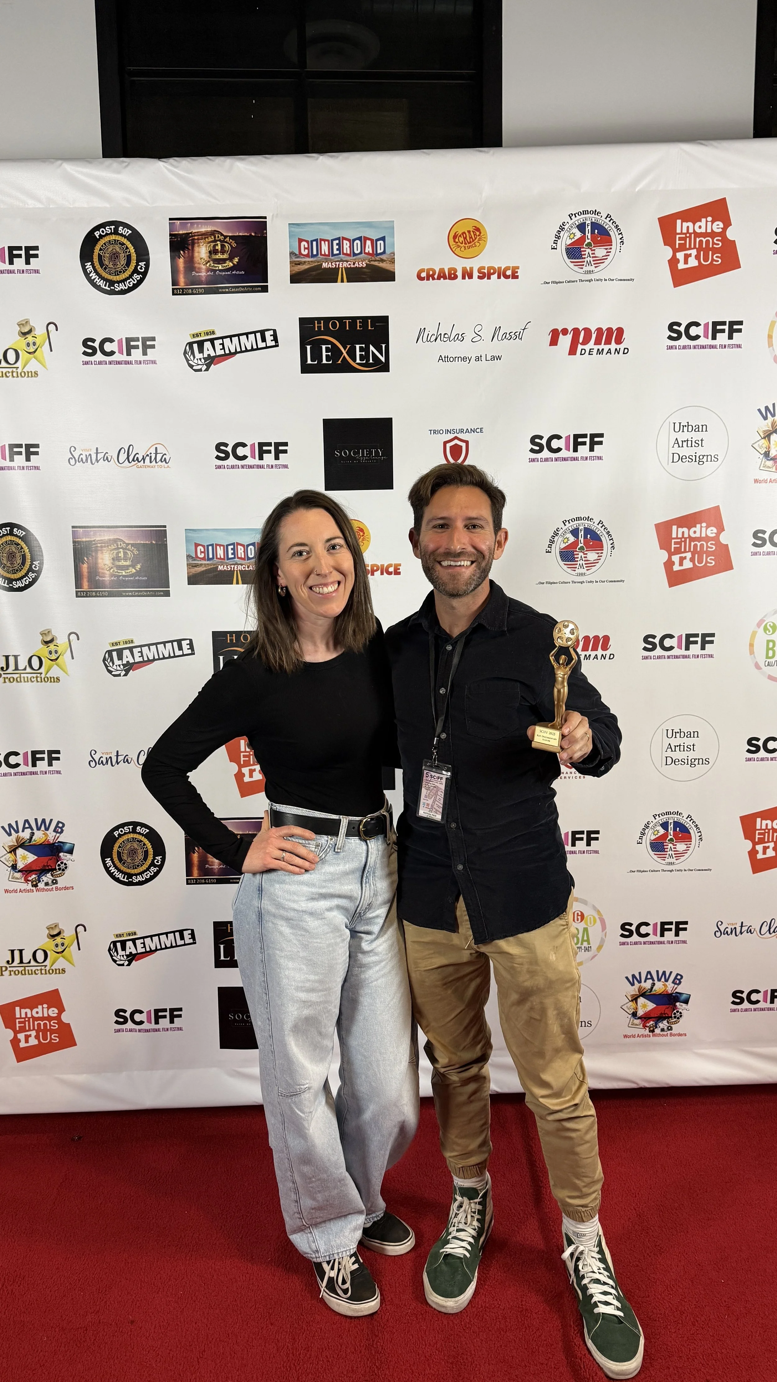 A man and woman stand together at an event, smiling. The man holds a trophy. The background has logos of various sponsors and the event's branding on a step-and-repeat banner.