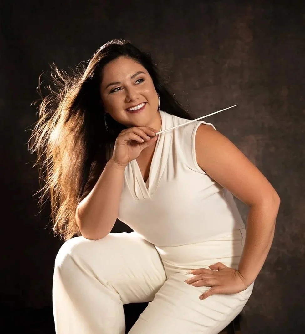 Danielle poses in an active shot, holding a conductor's baton in her one hand with the other on her hip. Her long brown hair is flowing and back lit. She wears a jumpsuit.