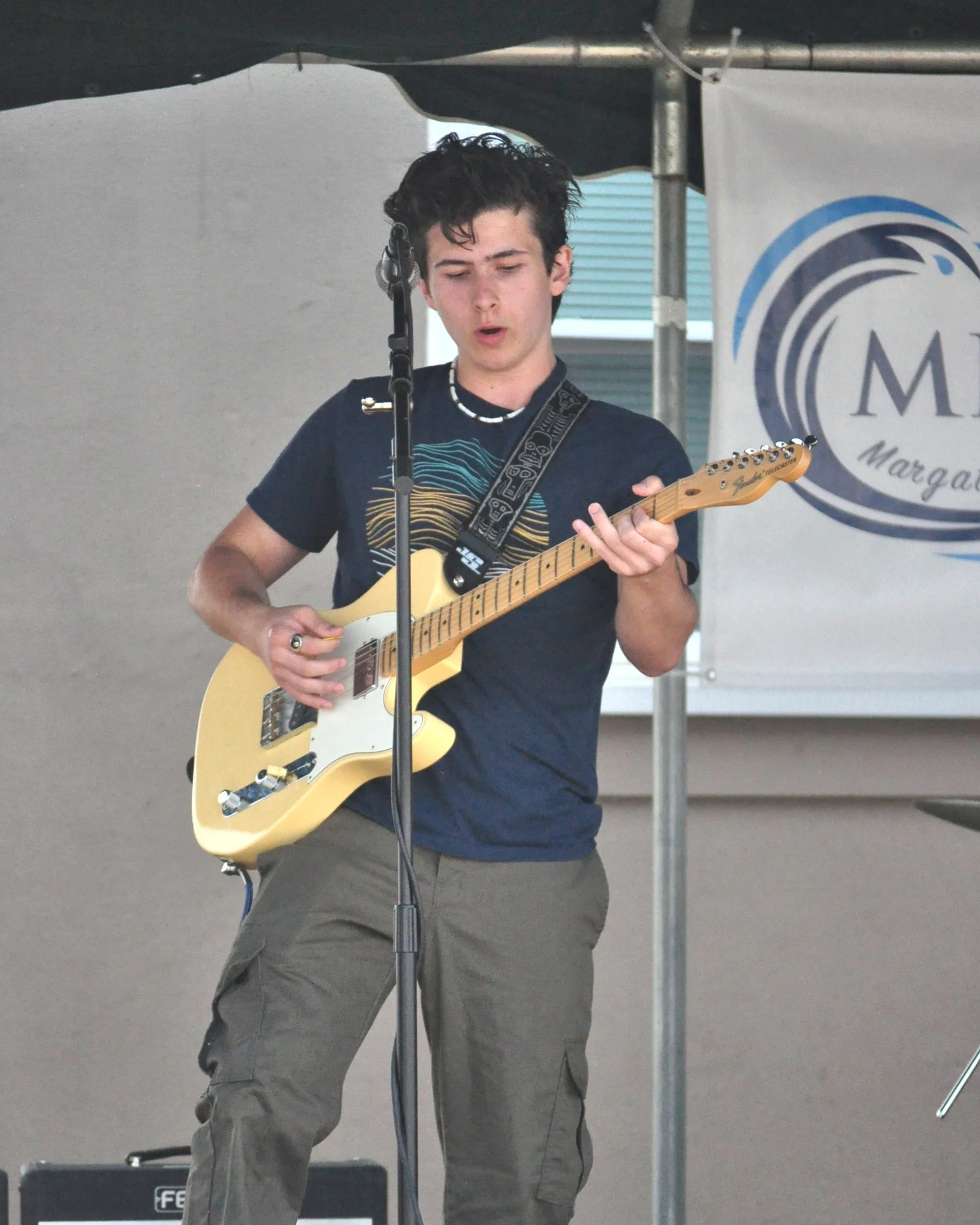 A young man with dark, curly hair playing an electric guitar on an outdoor stage, with a banner that reads 'MBA - Margate Has M' in the background.