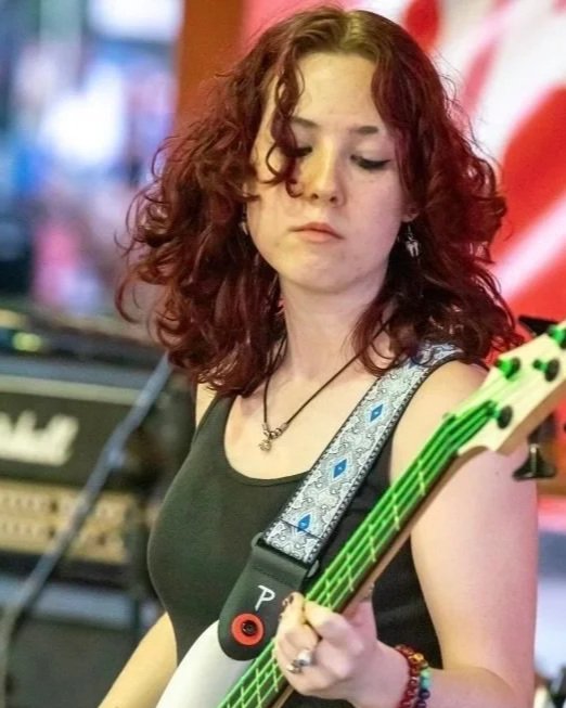 A young woman with curly red hair playing an electric guitar on stage, with musical equipment in the background.