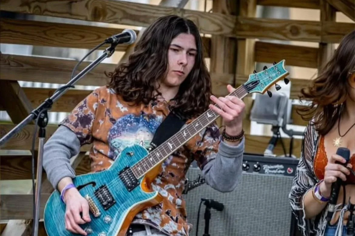A young man with long hair playing a blue electric guitar during a live outdoor performance, with musical equipment and a wooden stage in the background.