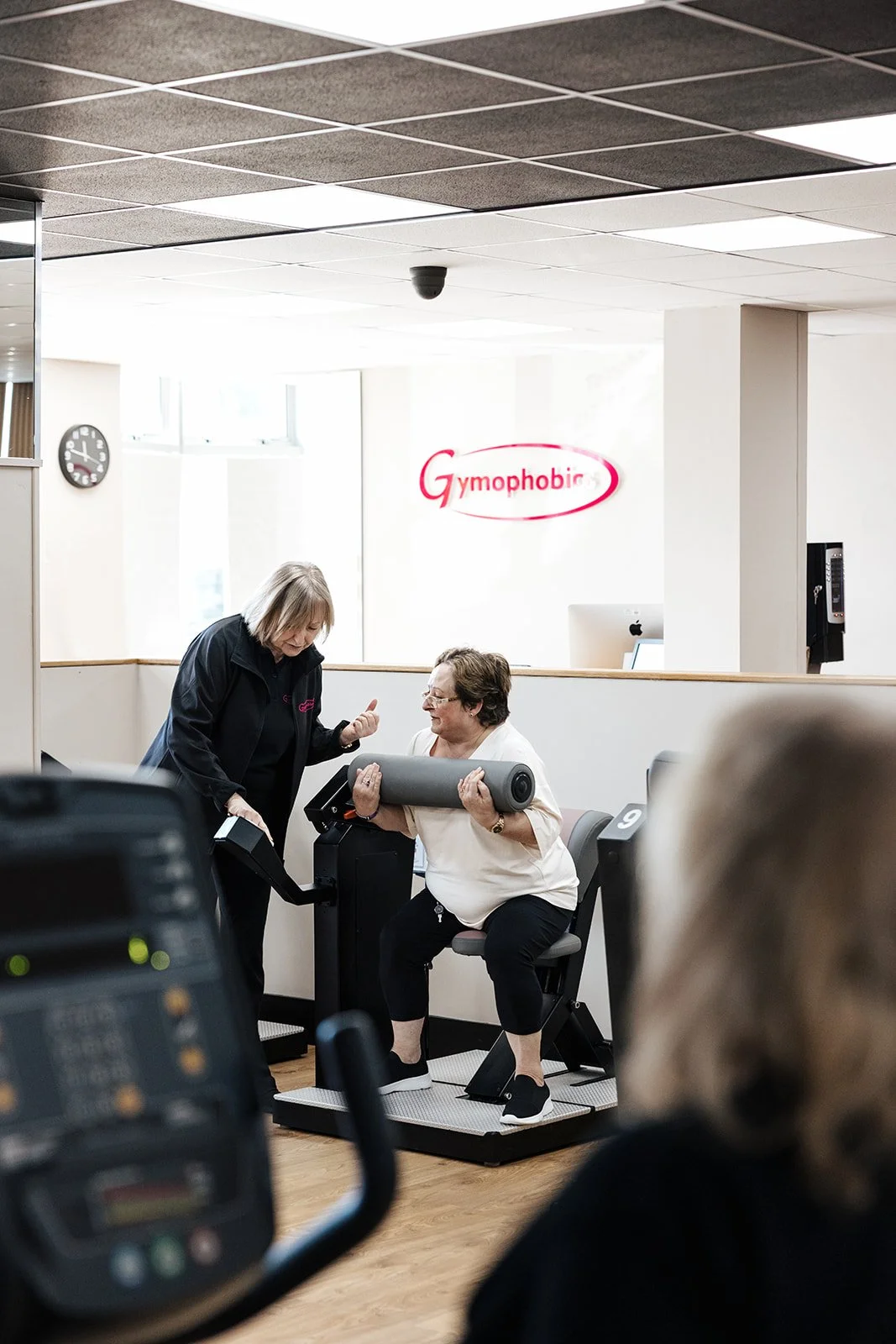 Gym instructor helping an older woman use a seated strength-training machine in a gym