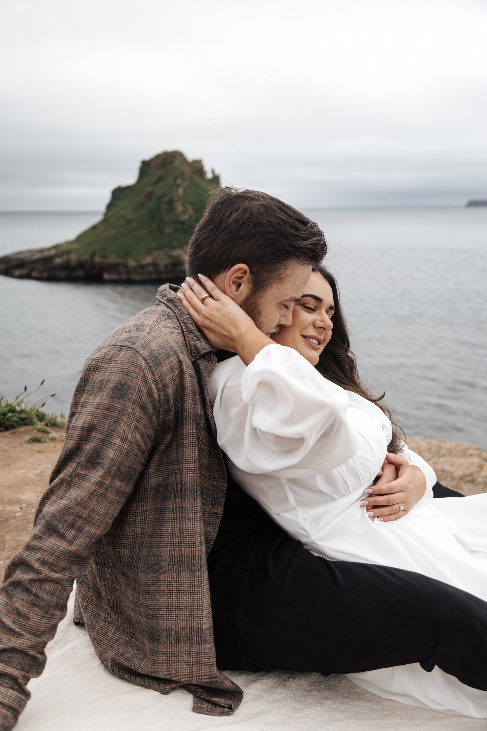 A couple embraces near the ocean with an island in the background, the woman sitting and the man leaning into her with his eyes closed, both appearing happy and content.