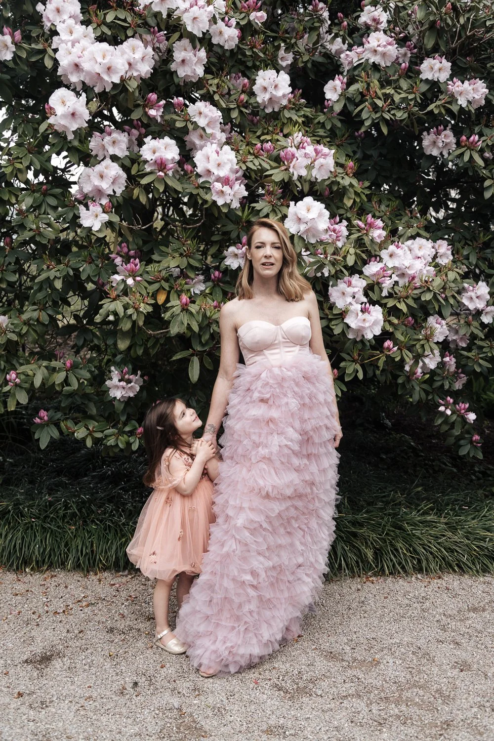 A woman and a young girl holding hands in front of a large blooming pink azalea bush. The woman is wearing a strapless, pink ruffled gown, and the girl is wearing a peach-colored dress with floral embroidery.