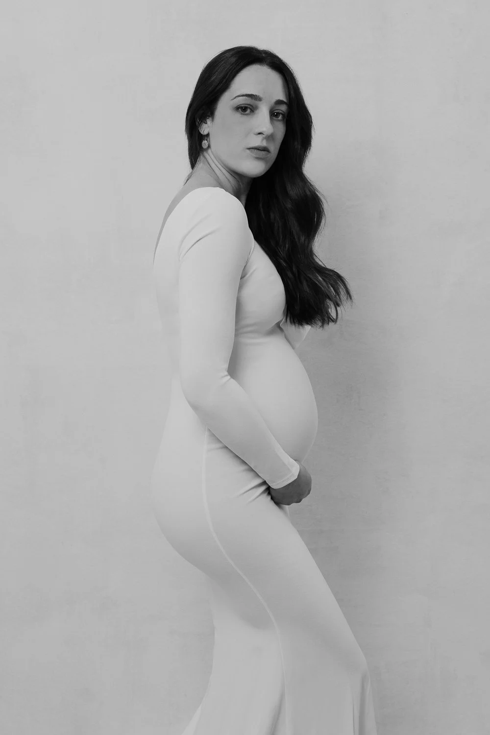Black and white photo of a pregnant woman in a fitted long sleeve dress, with long wavy hair, looking at the camera against a plain wall.