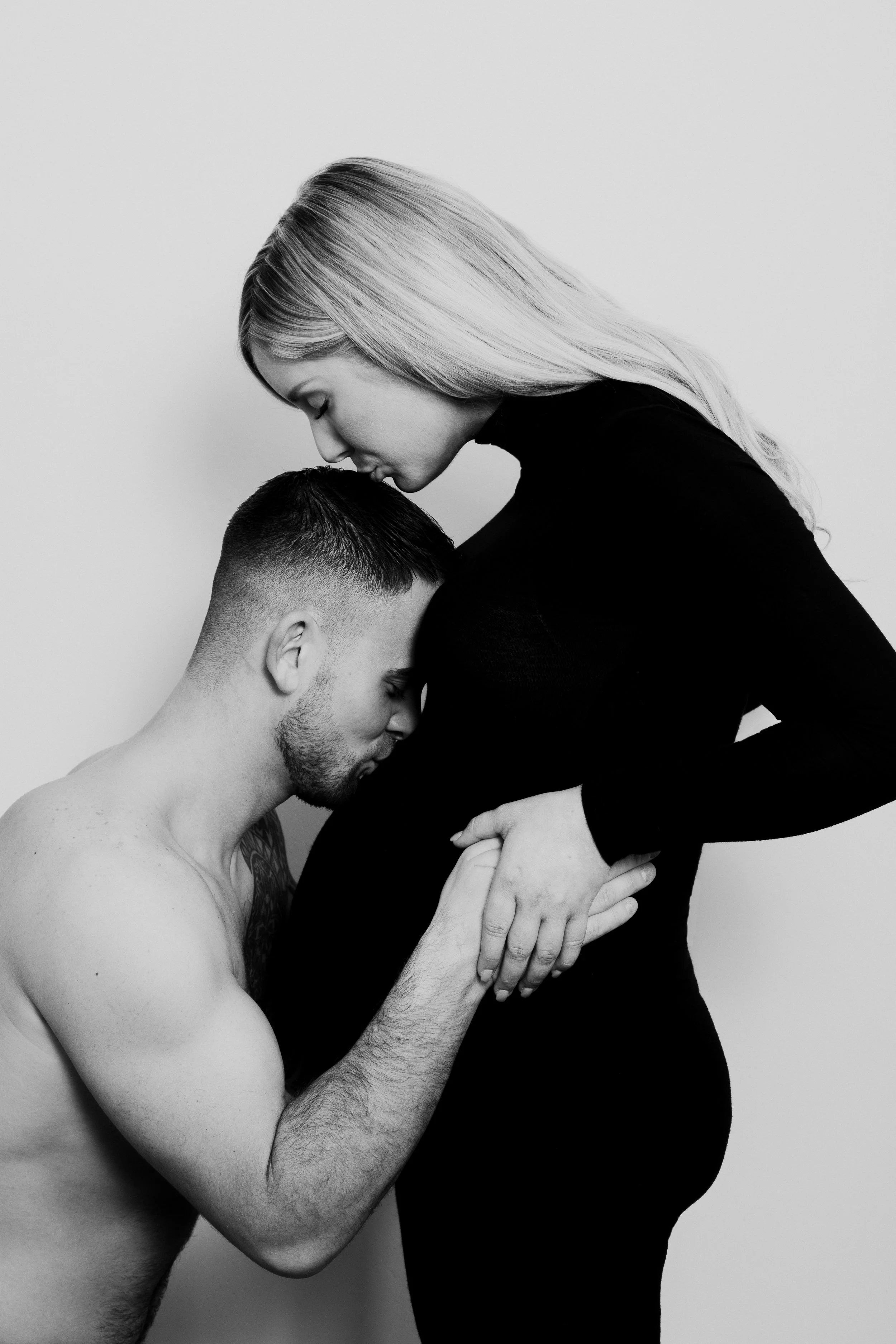 Black and white photograph of a shirtless man with closed eyes, holding a pregnant woman's belly, while she leans her forehead against his and touches her belly with her hands.