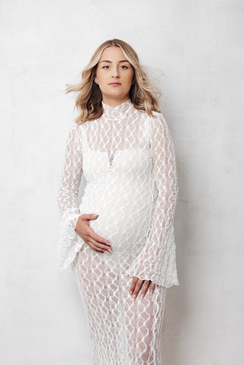 a pregnant woman with long wavy hair, posing with one hand on her belly, against a plain background.