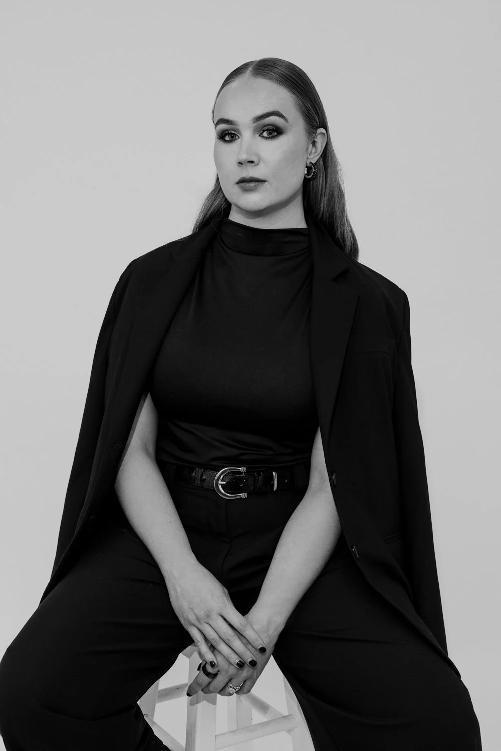 Black and white portrait of a young woman with long hair, wearing a blazer over a turtleneck, sitting on a stool with her hands clasped in front of her.