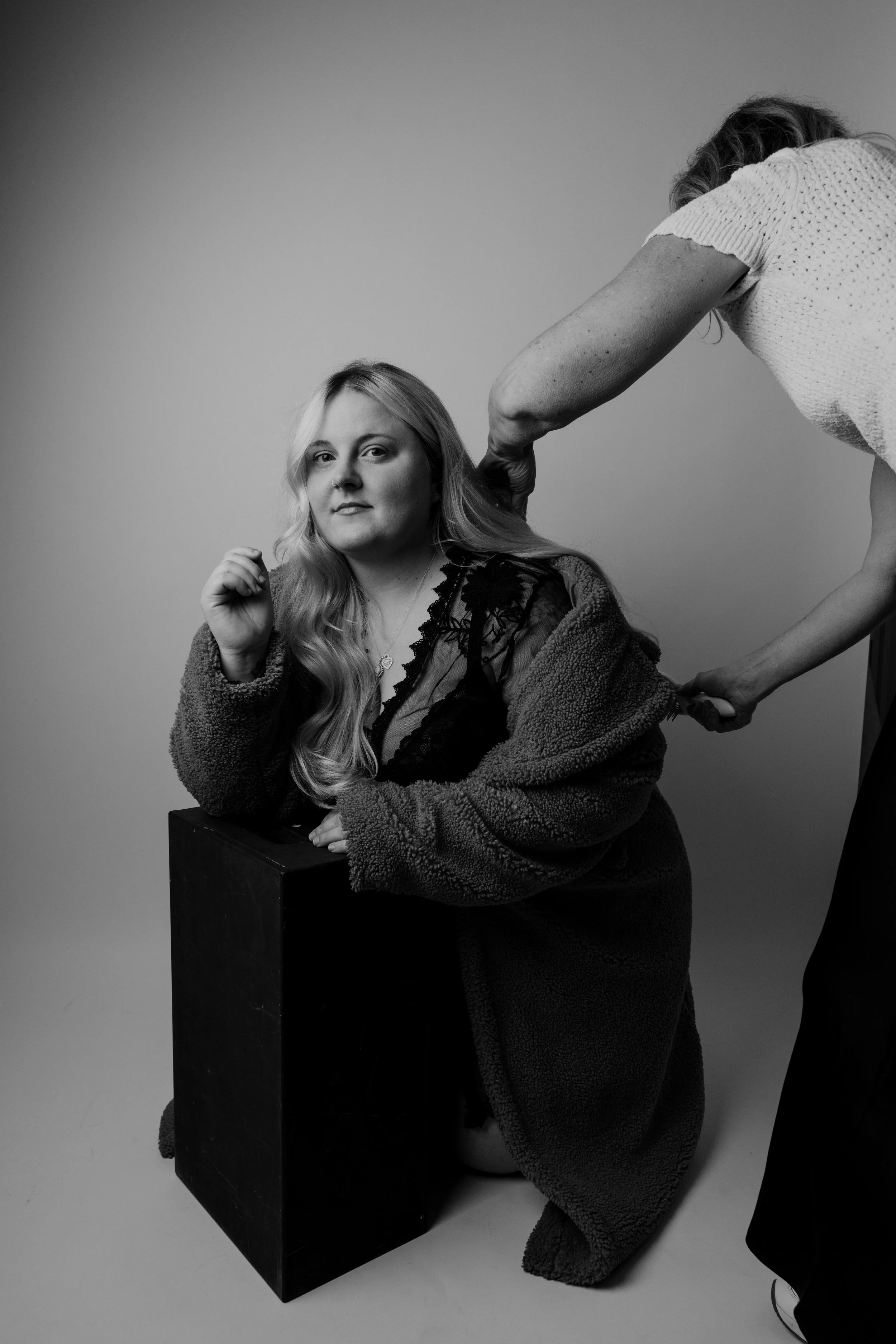 A woman with long, wavy hair is sitting on a box, wearing a dark lace dress and a cozy jacket, with a serious facial expression. Another person, partially visible, is adjusting the woman's hair.