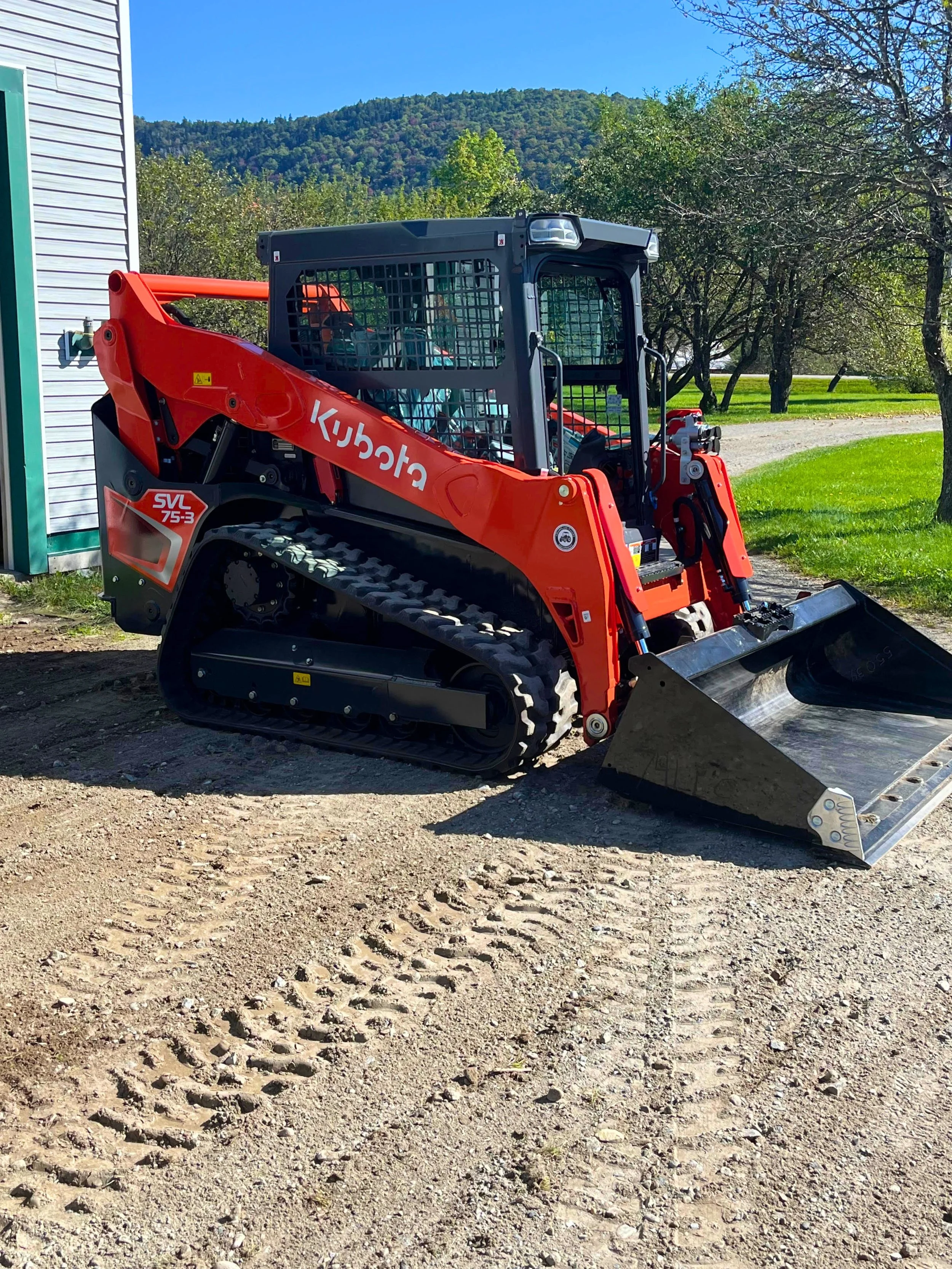 A Kubota SVL75-3 compact track loader parked on a gravel surface with tire tracks. Trees and hills are visible in the background.