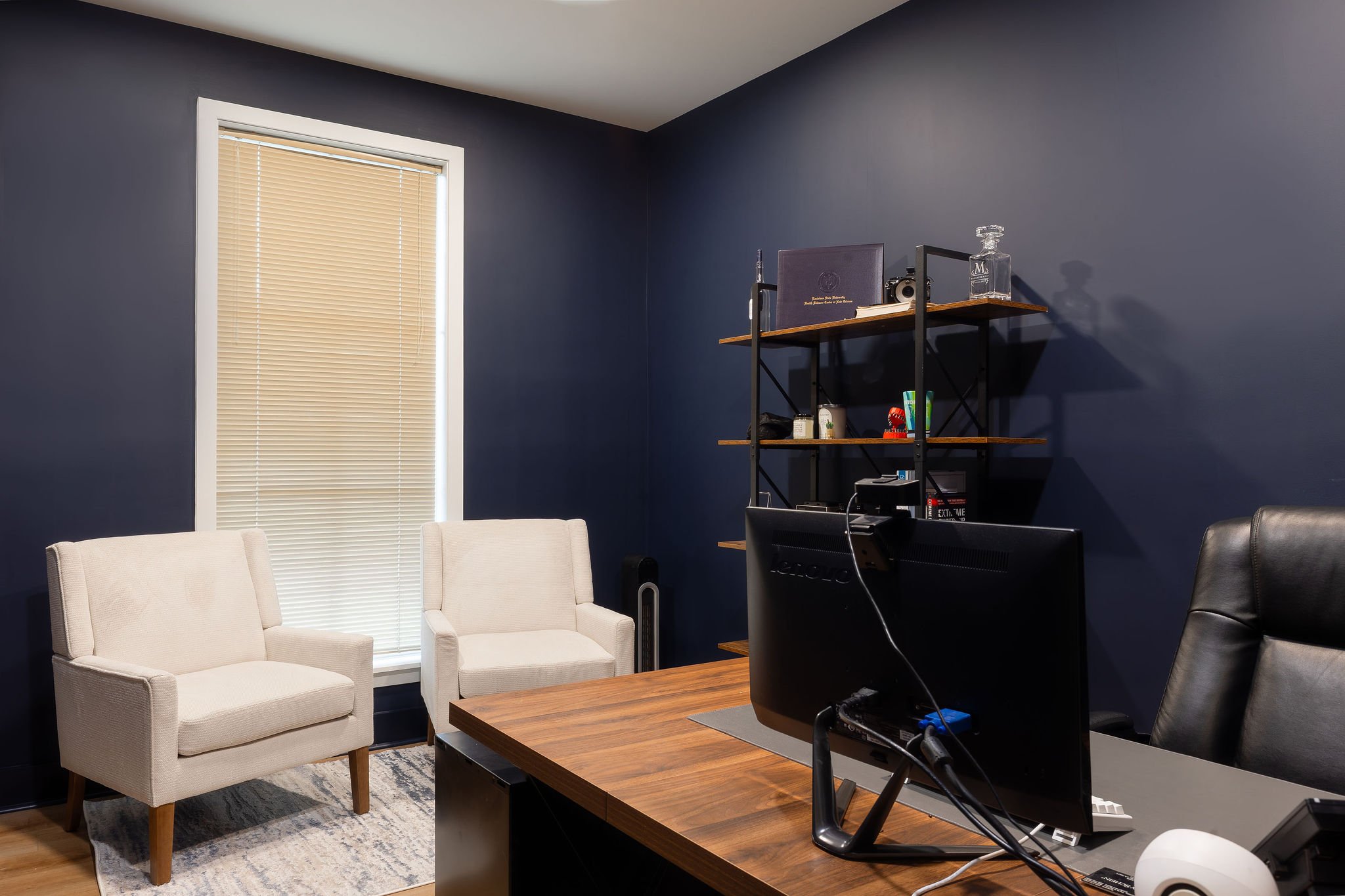 Office room with dark blue walls, a window with beige blinds, a wooden desk with a black office chair, a computer monitor, a black shelving unit with books and decorative items, and two white armchairs by the window.