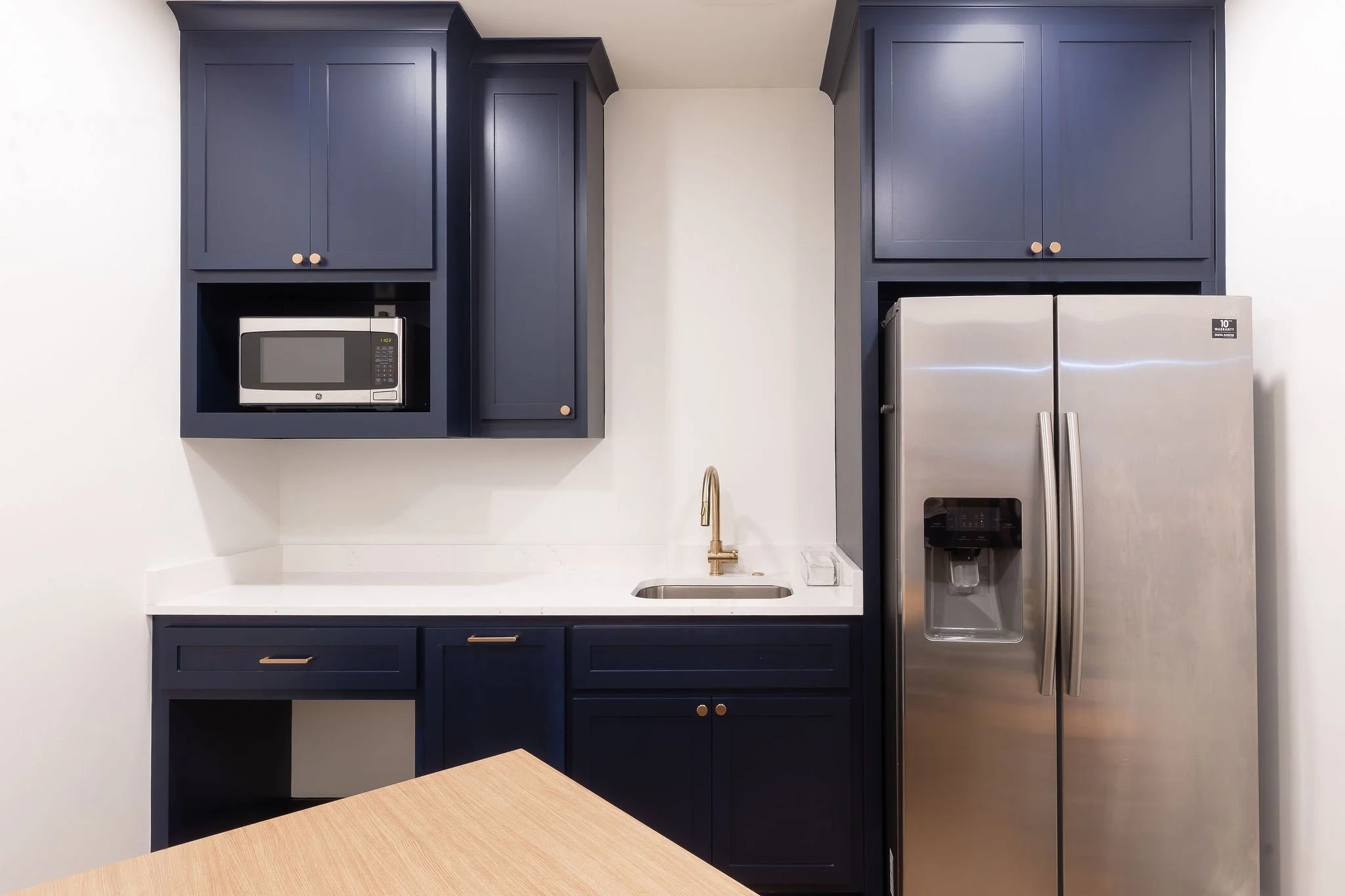 Kitchen with navy blue cabinets, a white countertop, a microwave in a cabinet, a double-door stainless steel refrigerator with an ice and water dispenser, a small sink, and a brass faucet.