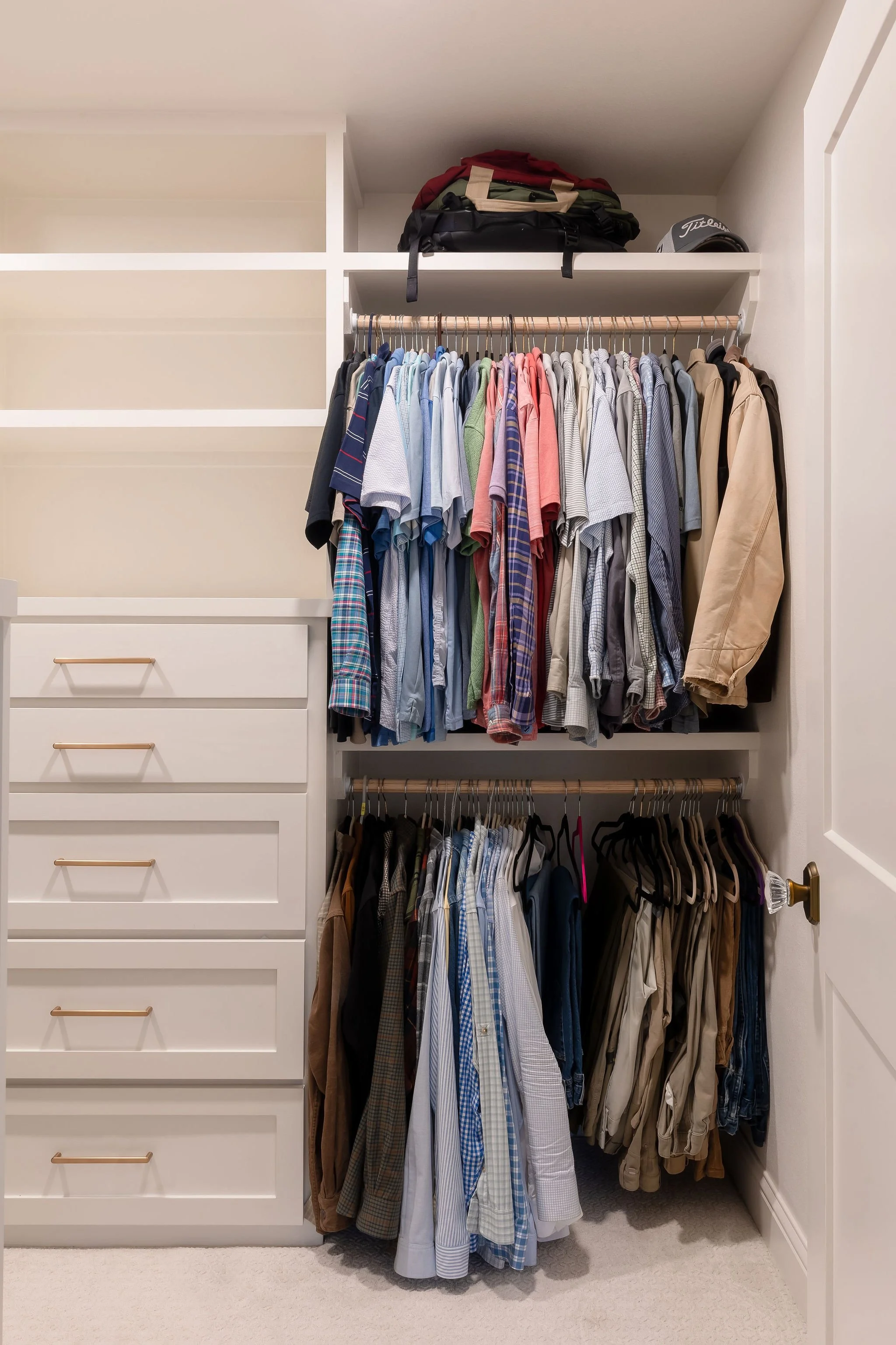 Inside a closet with hanging shirts on two rods, stacked on top of each other. The upper rod has mostly short-sleeved shirts, while the lower rod has longer shirts and pants. There are also shelves above and a white chest of drawers to the left.