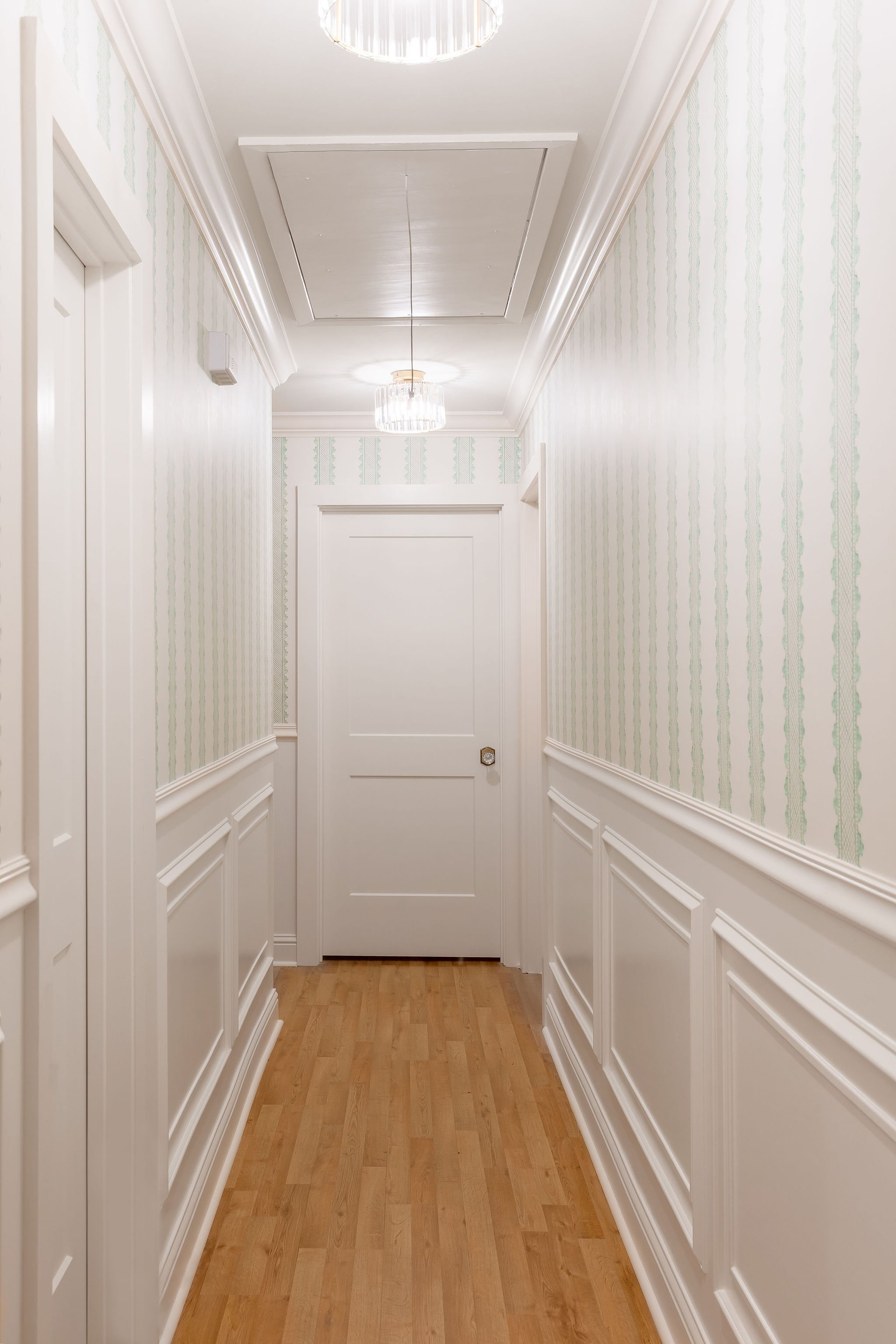 Hallway with white-paneled walls, patterned wallpaper, wooden floor, and ceiling lights.