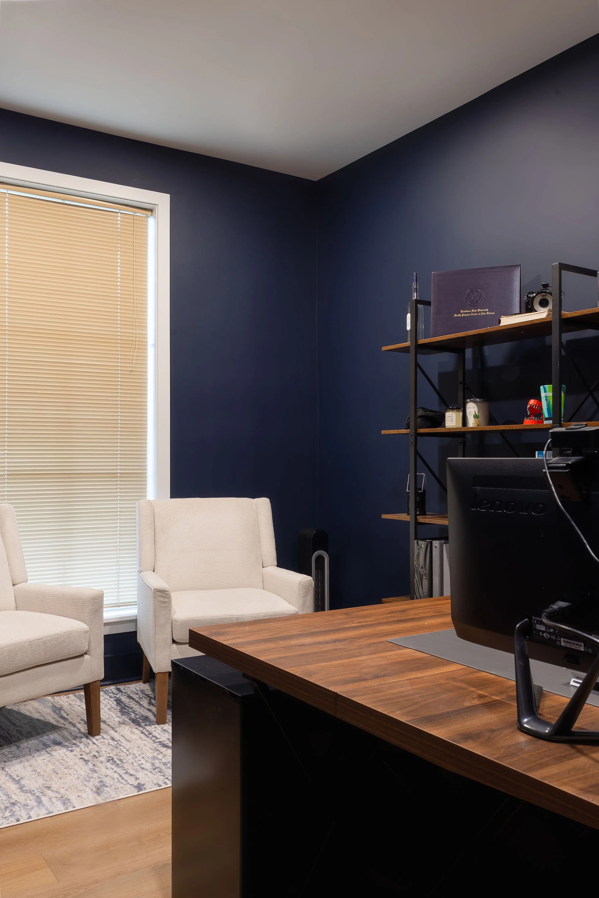An office space with a large wooden desk, two beige armchairs, a window with beige blinds, a black metal shelf with wooden shelves, and a computer monitor on the desk.