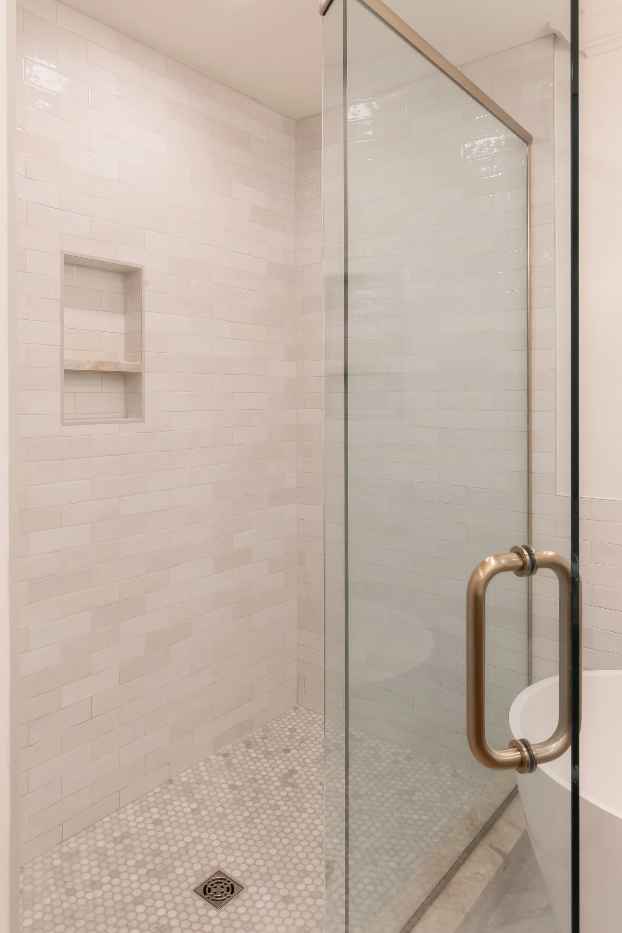 Empty shower with white subway tile walls, a built-in niche with a small shelf, a glass door, a handle, and a floor drain with small round tiles.