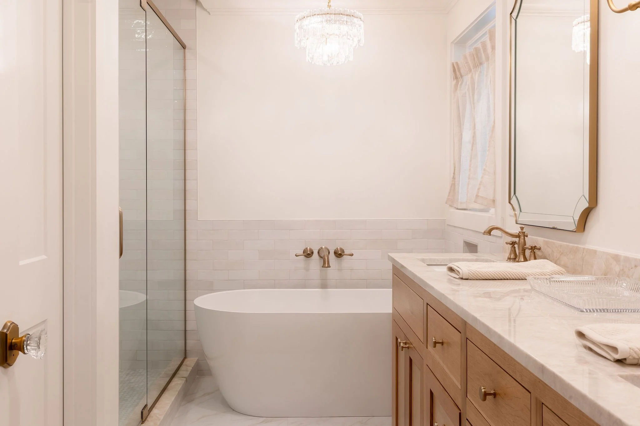 Modern bathroom with white bathtub, marble countertop, gold fixtures, wooden vanity, and a glass shower door