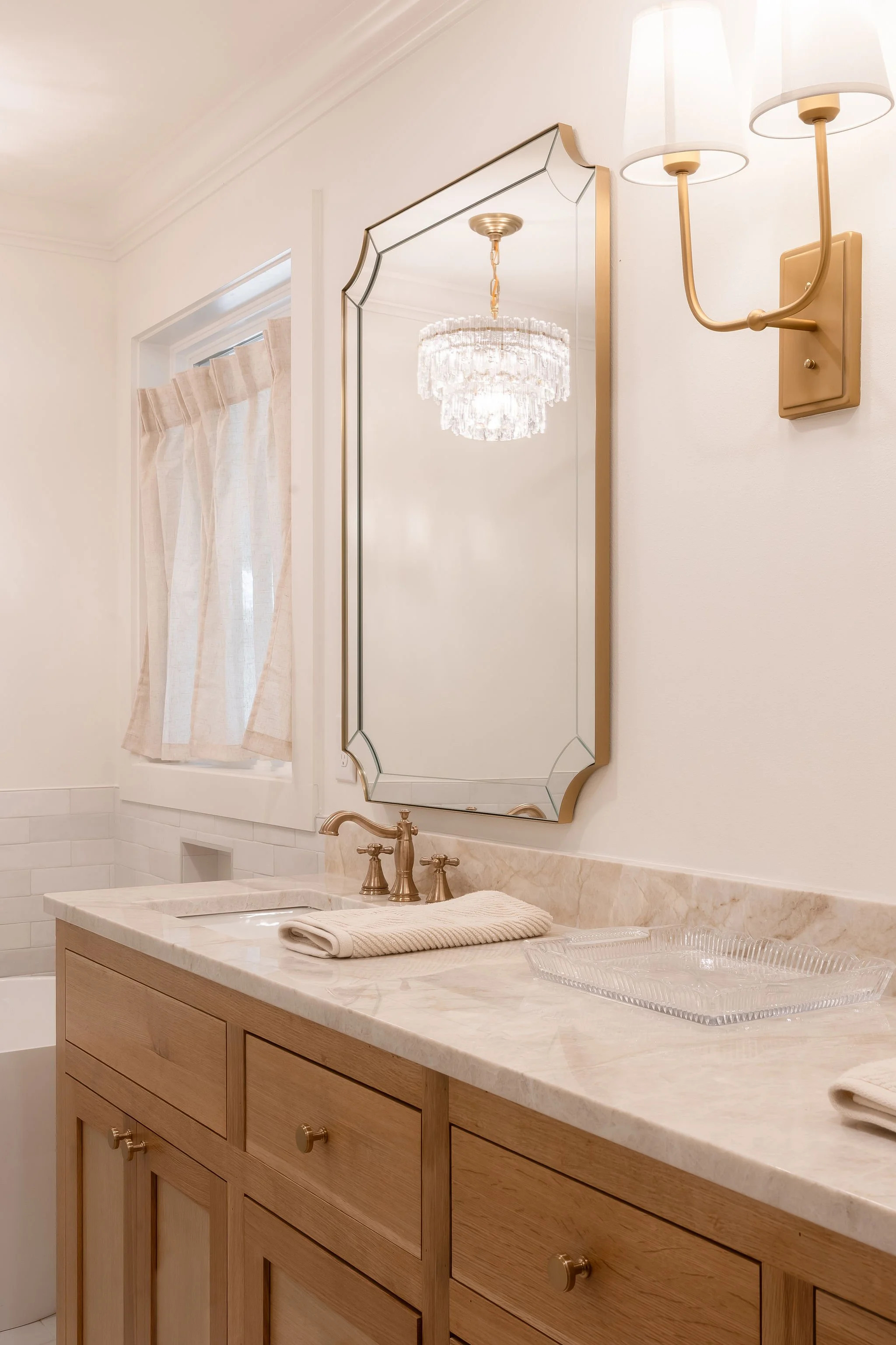 A bathroom with a marble countertop, a wooden cabinet, a mirror, and a wall-mounted lamp with white shades. A window with beige curtains and a chandelier reflected in the mirror.