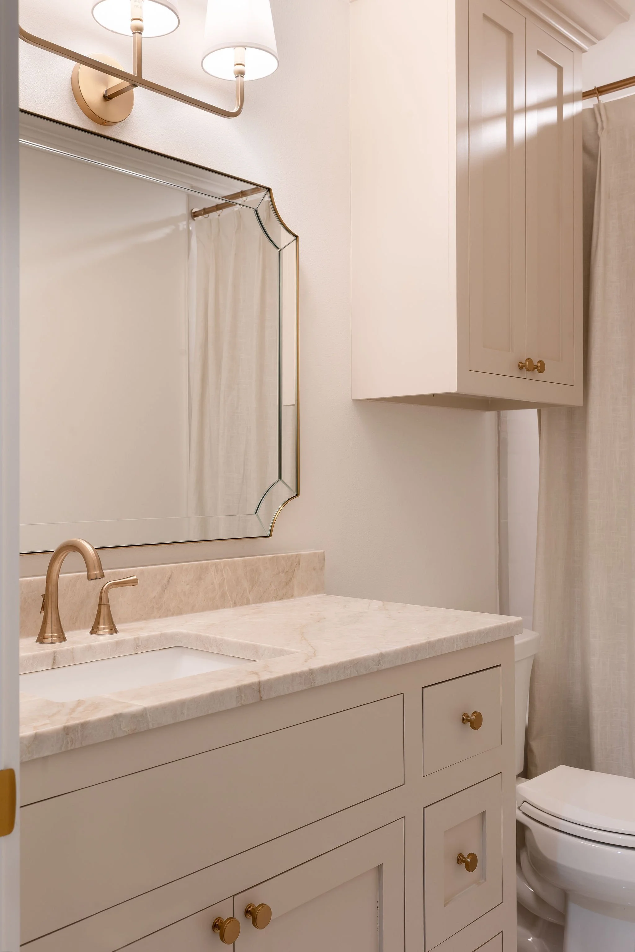 A bathroom with a marble countertop, a sink with a gold faucet, a large mirror with a decorative gold frame, a wall-mounted light fixture with two bulbs, a white cabinet, and a toilet with a beige curtain.